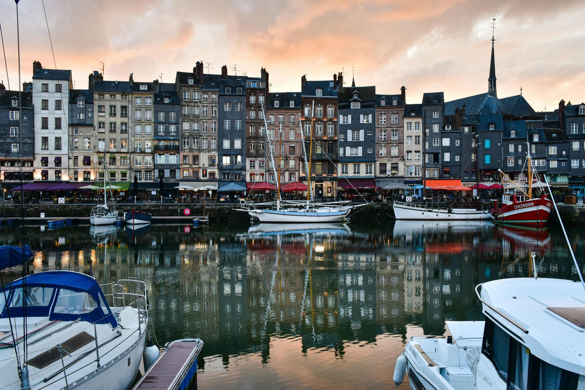 A row of boats are docked in a harbor with buildings in the background in Normandy, France.