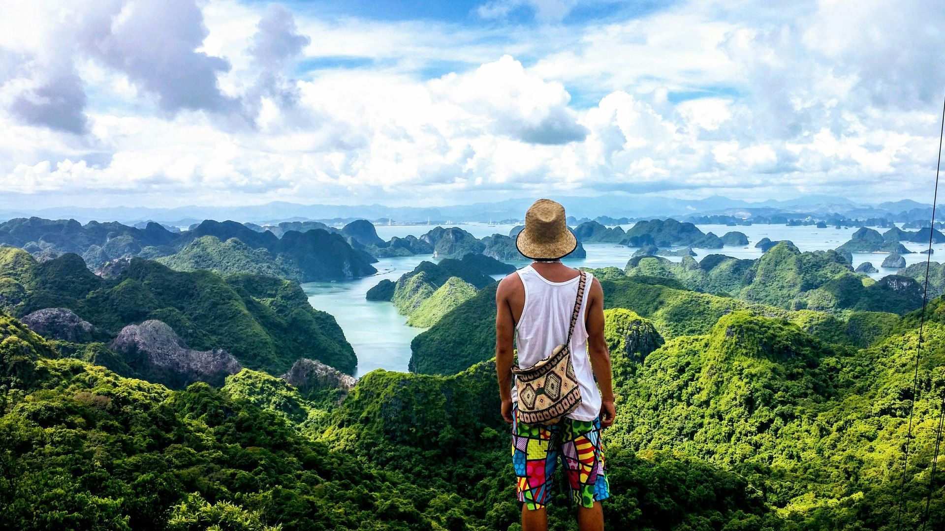 A man in a hat is standing on top of a mountain overlooking Hạ Long Bay in Vietnam.