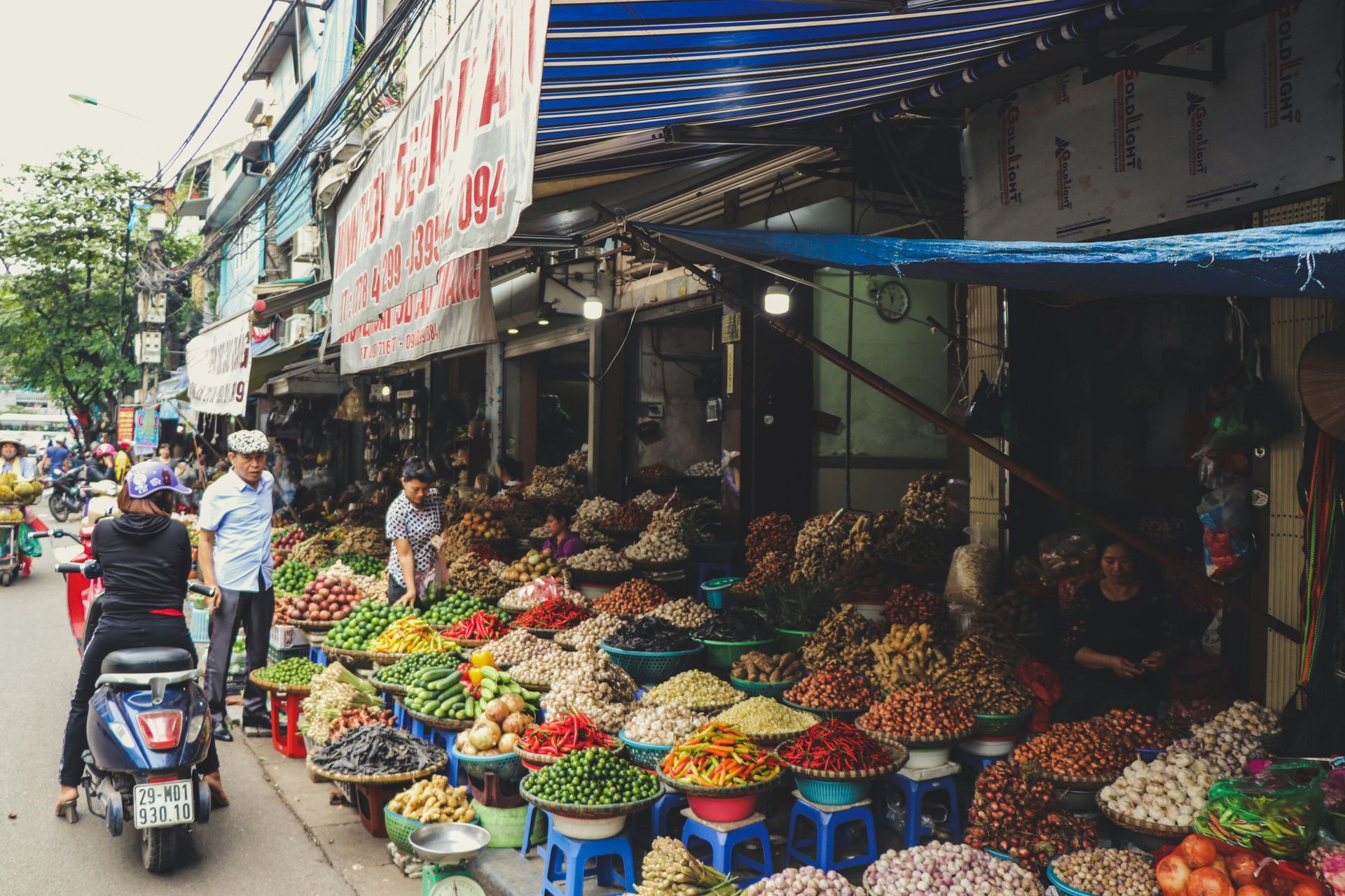 A man is riding a scooter down a street in front of a market filled with fruits and vegetables in Vietnam.