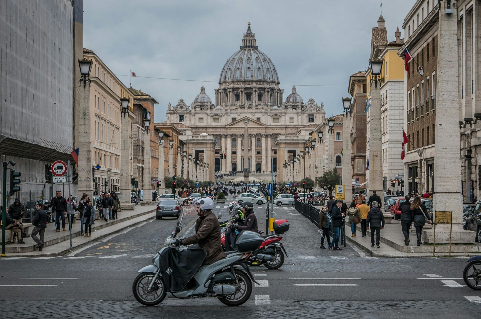 A man is riding a motorcycle down a street in front of a large building in Rome, Italy.