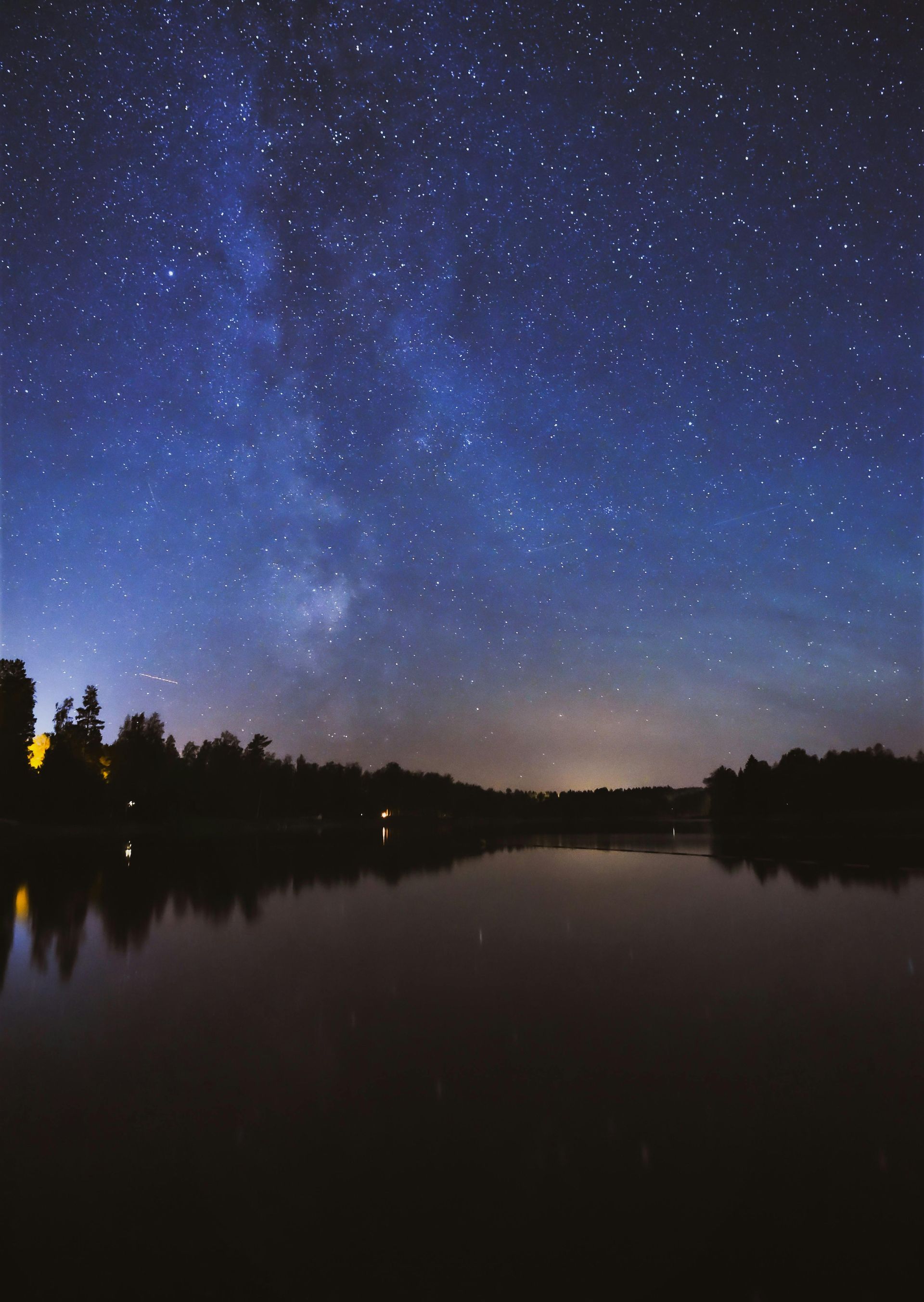 A starry night sky over a lake with trees in the foreground in Finland.