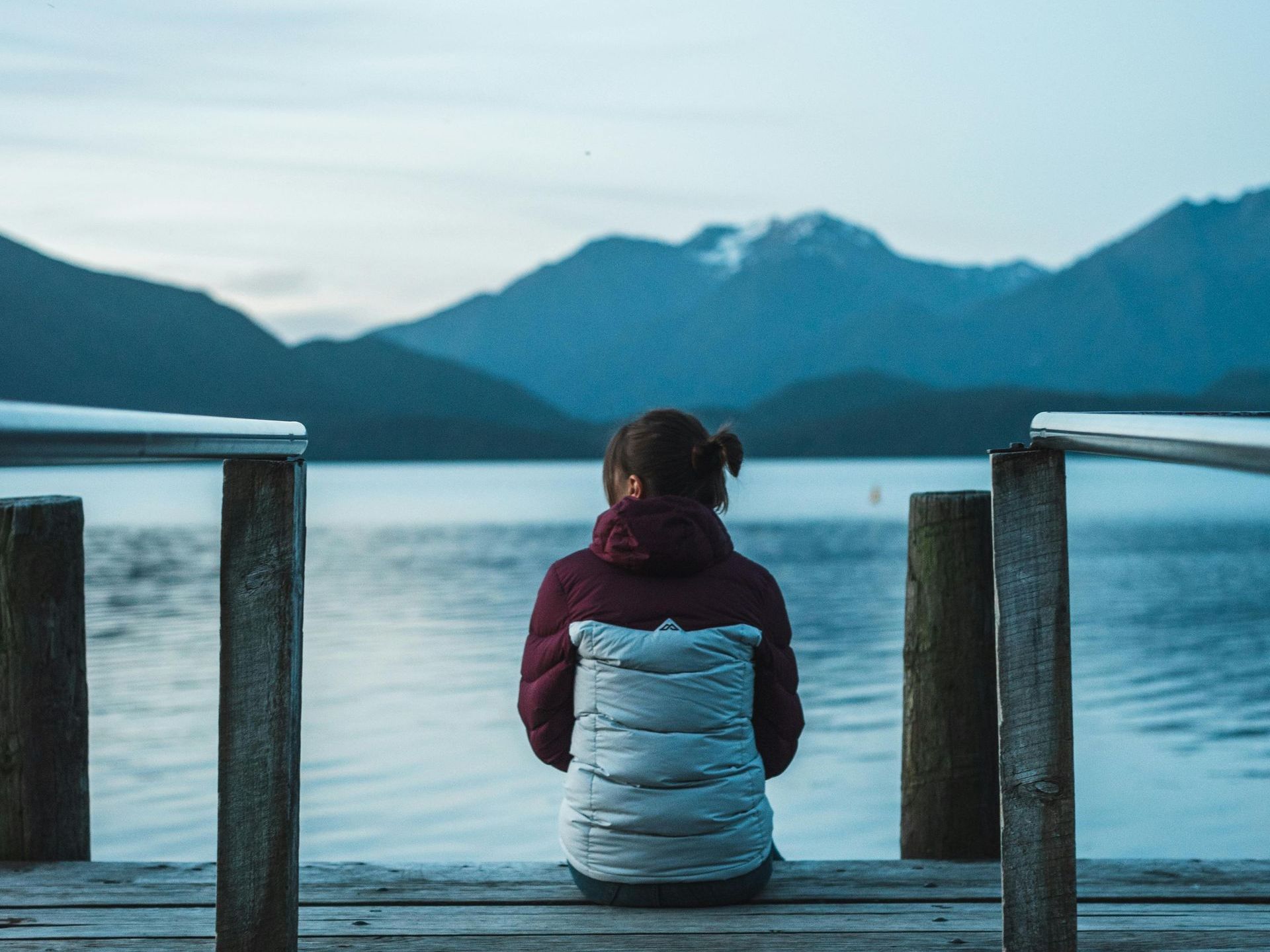 A woman is sitting on a dock looking at a lake with mountains in the background in New Zealand.