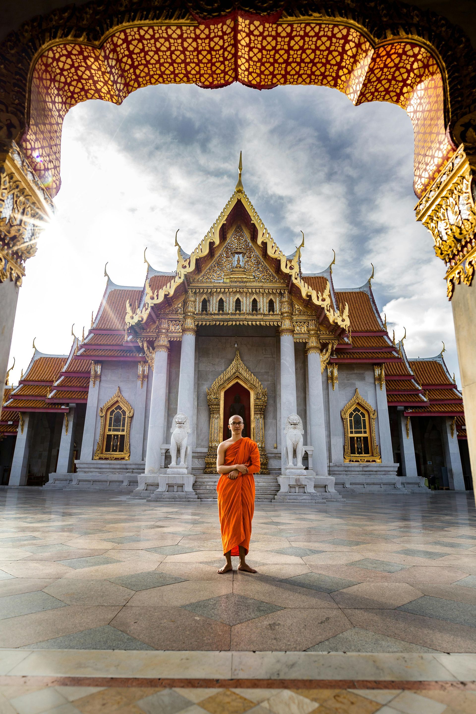 A monk in an orange robe is standing in front of a temple  in Bangkok, Thailand.