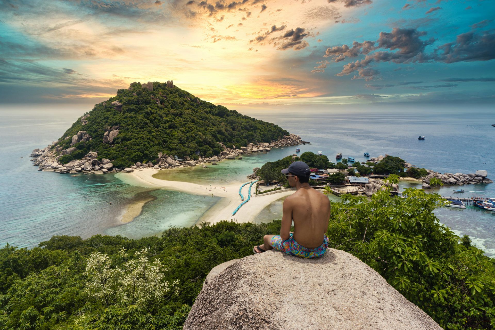 A man is sitting on top of a rock overlooking a small island in the ocean in Thailand.