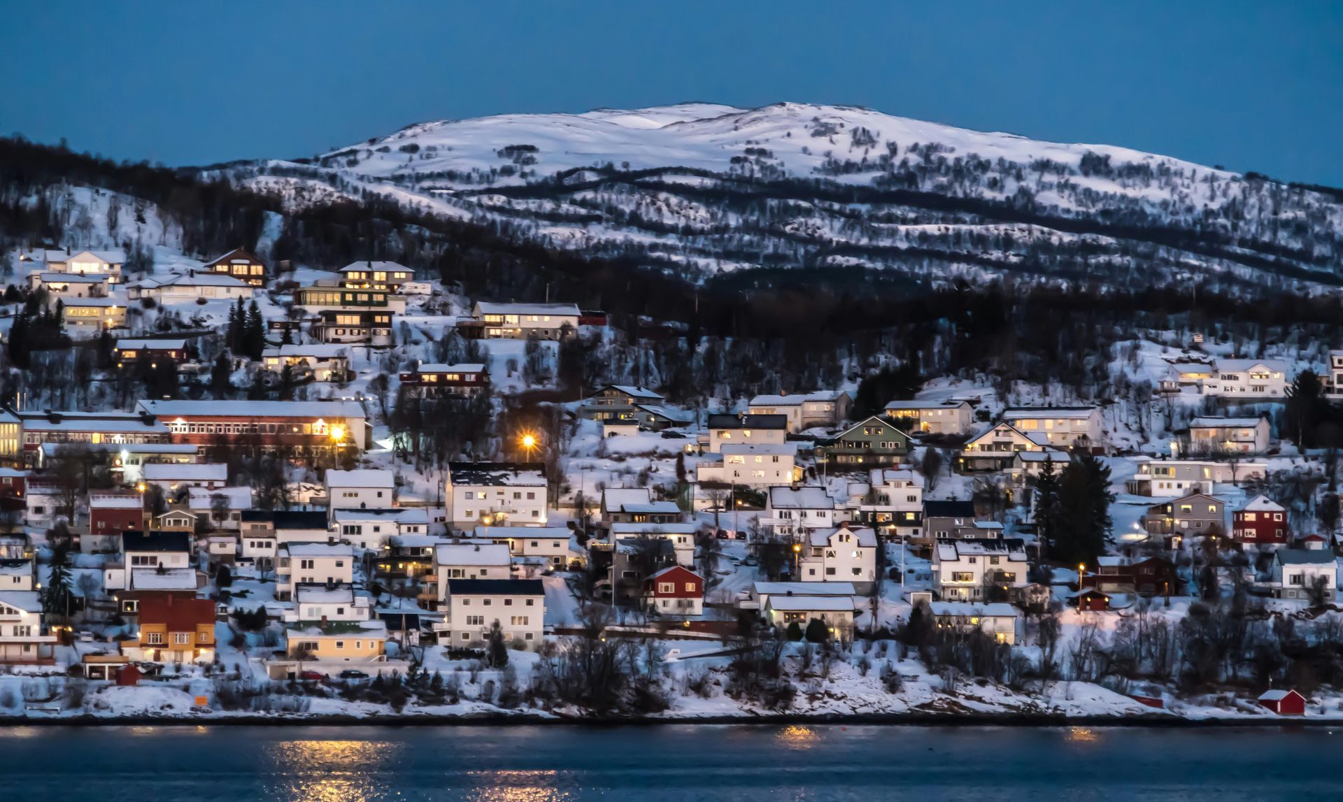 A small town is surrounded by snow covered mountains and a body of water  on the Norwegian Coast.