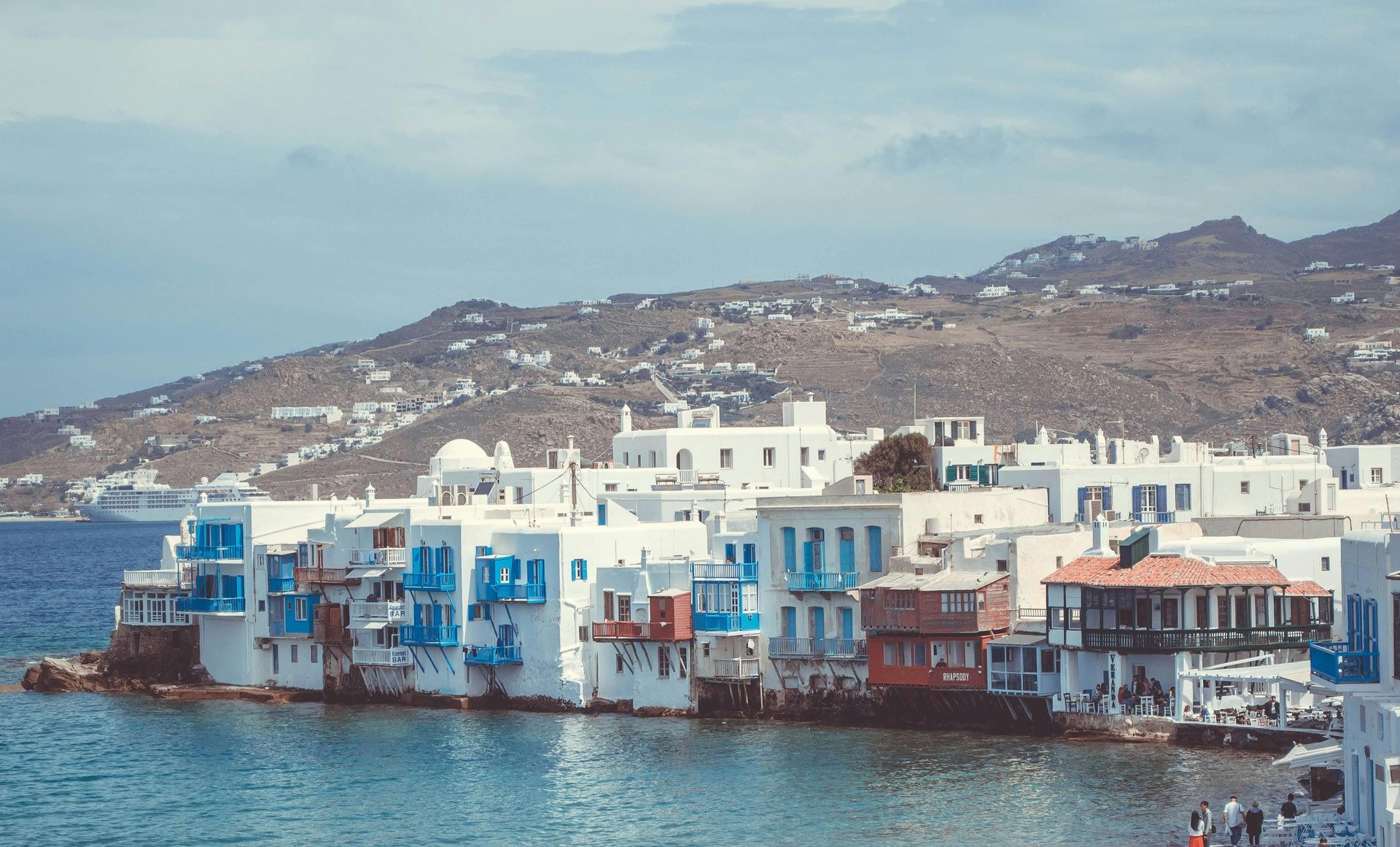 A row of white and blue buildings next to a body of water in Mykonos, Greece.