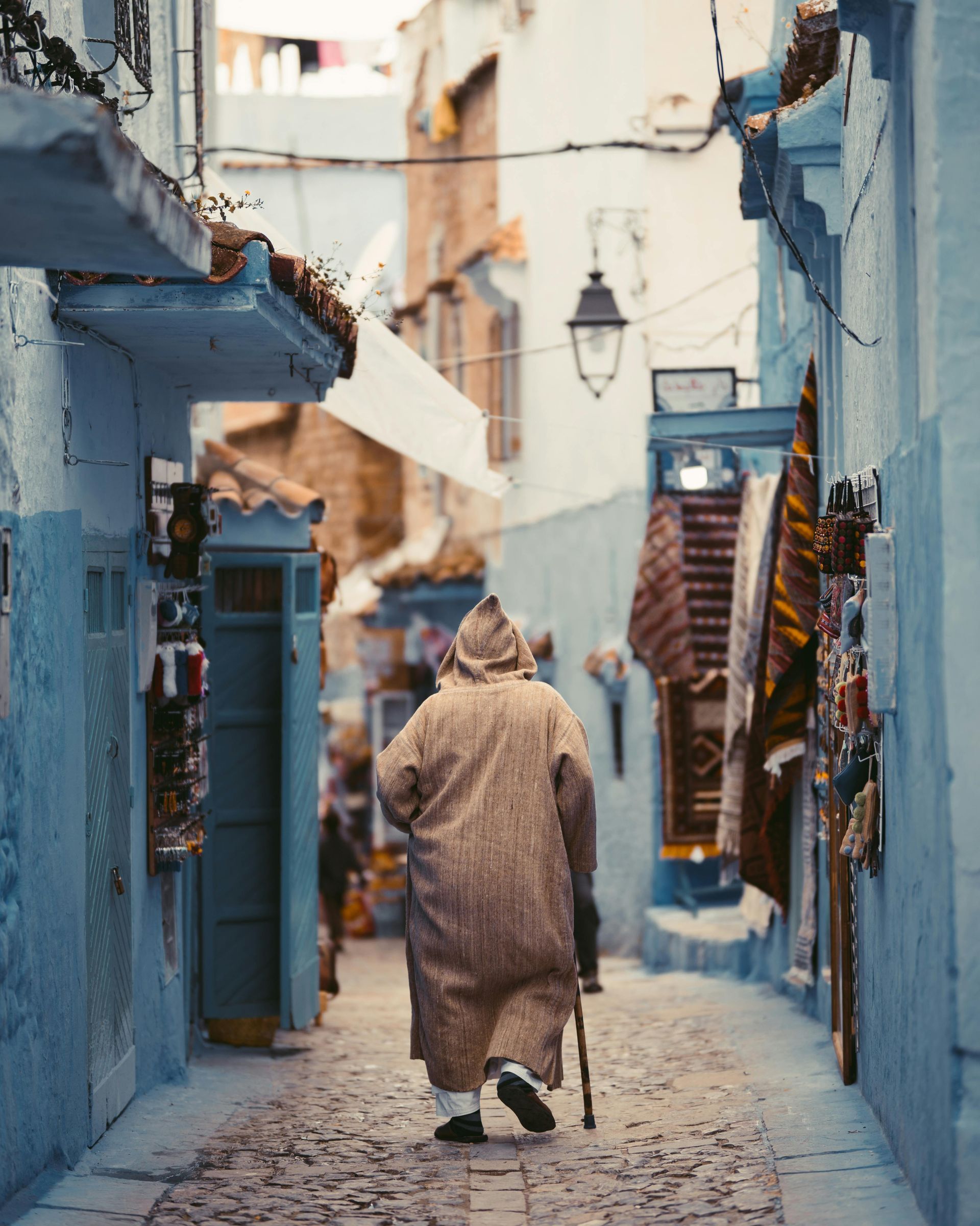 A man with a cane is walking down a cobblestone street in Morocco.