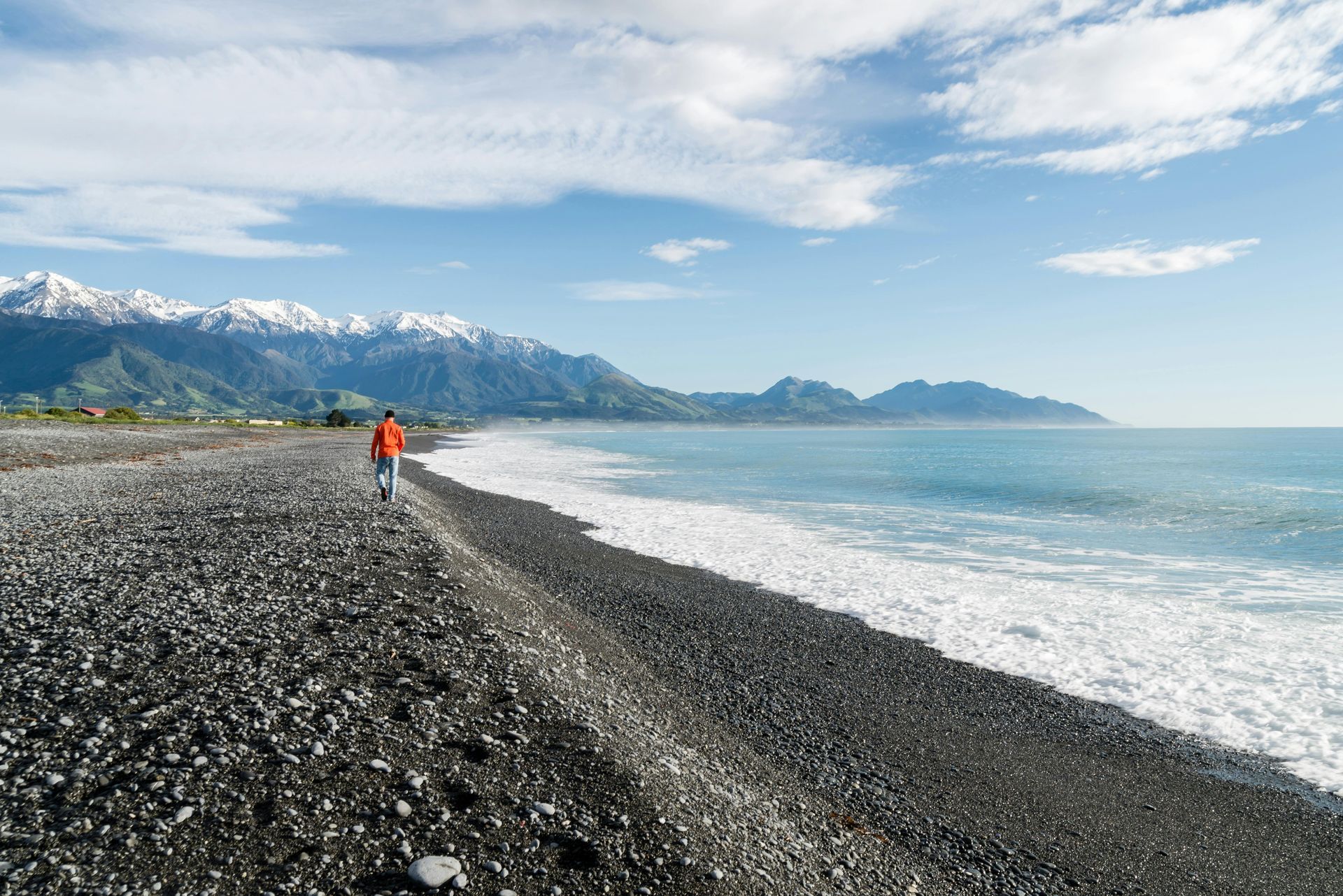 A man in an orange jacket is walking on a rocky beach near the ocean in Kaikōura, New Zealand.