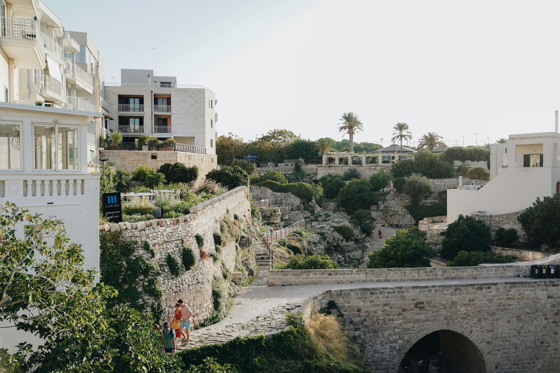 A couple of people are walking across a stone bridge in front of a building in Puglia, Italy.