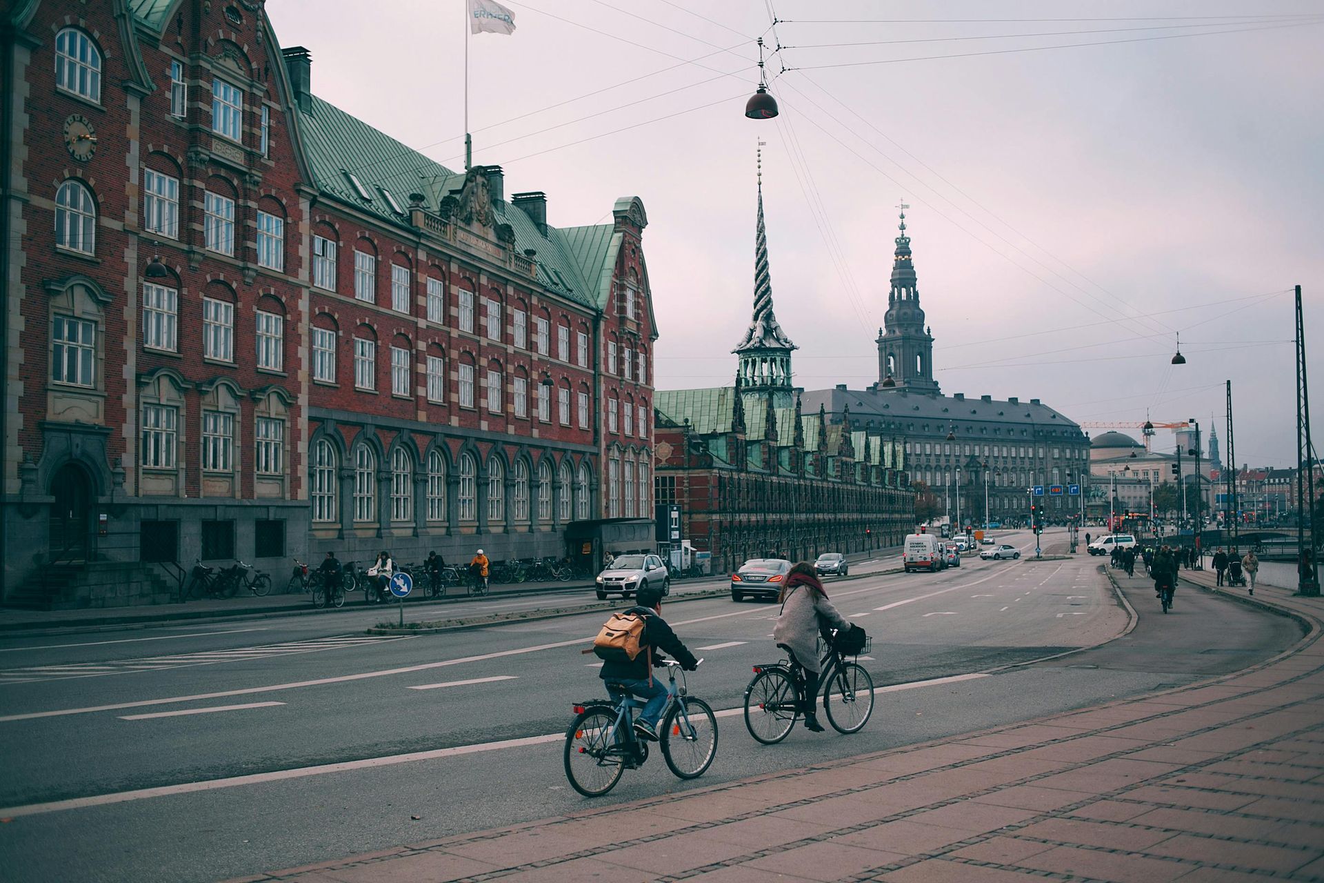 Two people are riding bicycles down a street of Copenhagen in front of a large building in Denmark.