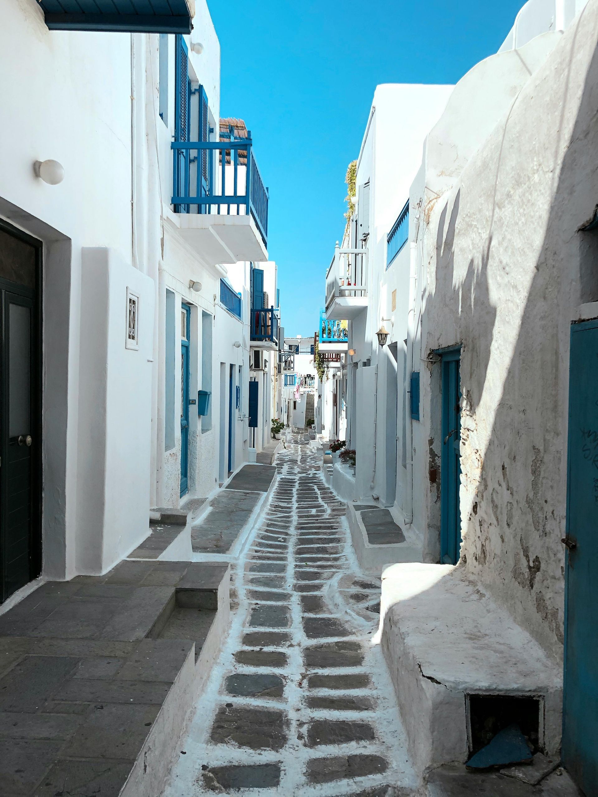 A narrow street with white buildings and blue balconies in Mykonos, Greece.