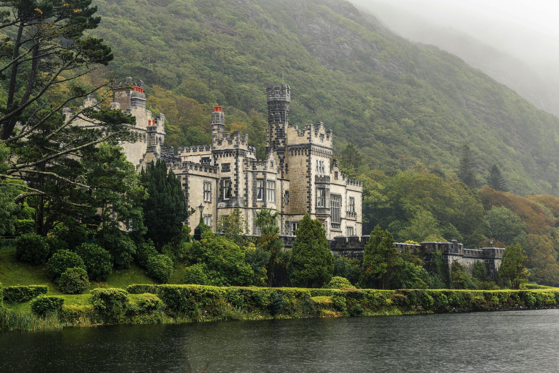 A large castle is sitting on top of a hill next to a lake in Kylemore, Ireland.