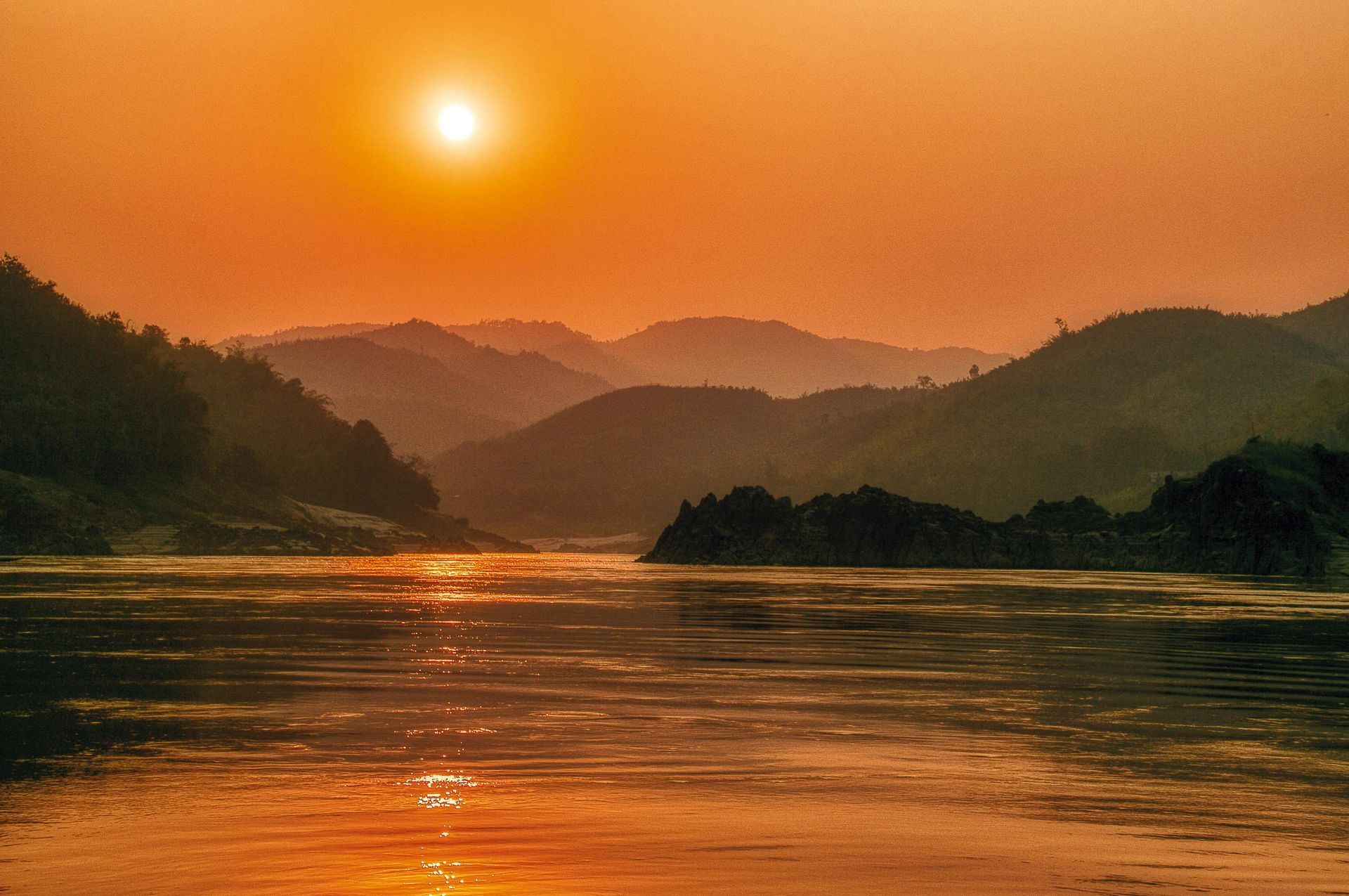 The sun is setting over the Mekong River with mountains in the background.