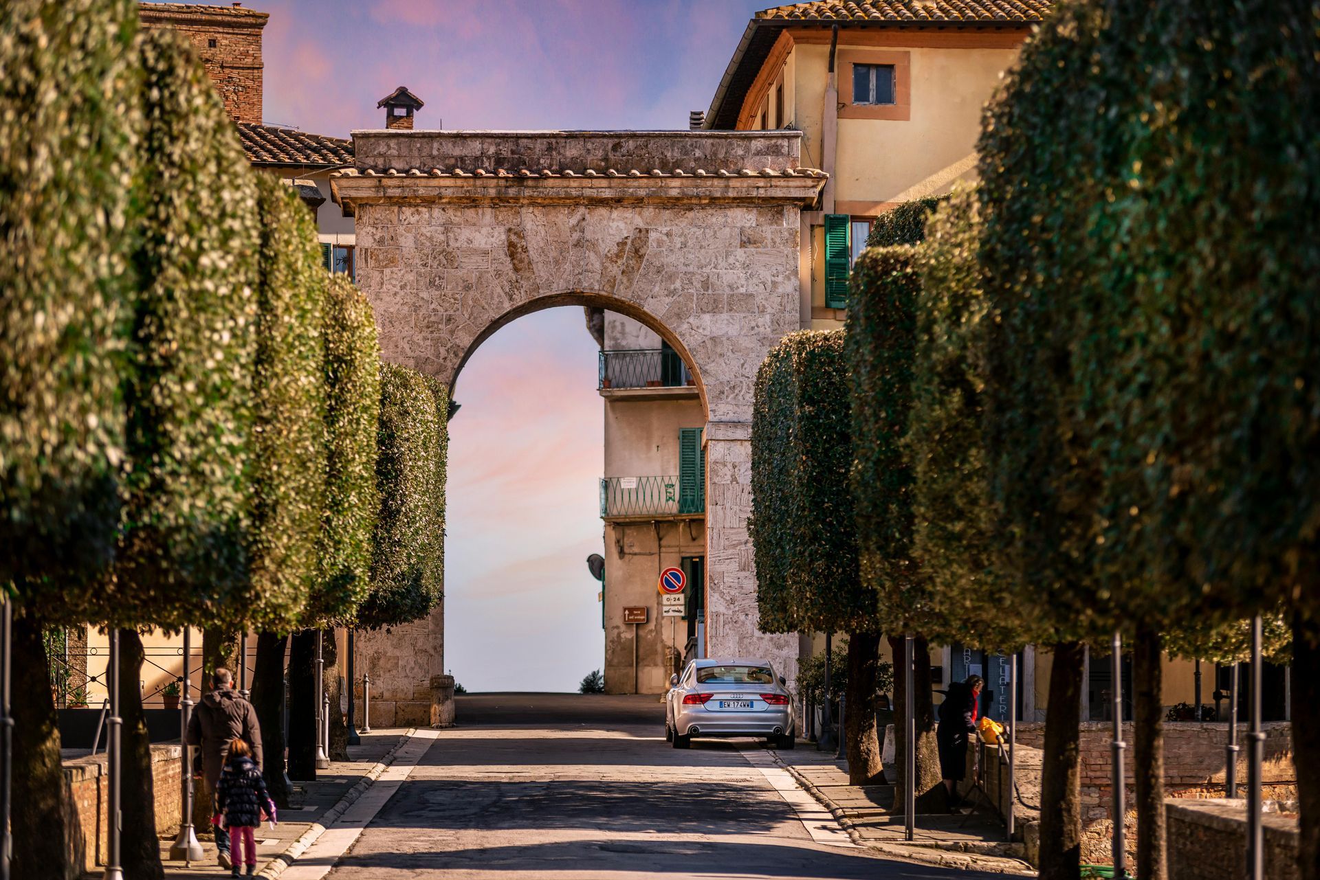 A car is parked under a stone archway surrounded by trees in Tuscany, Italy.