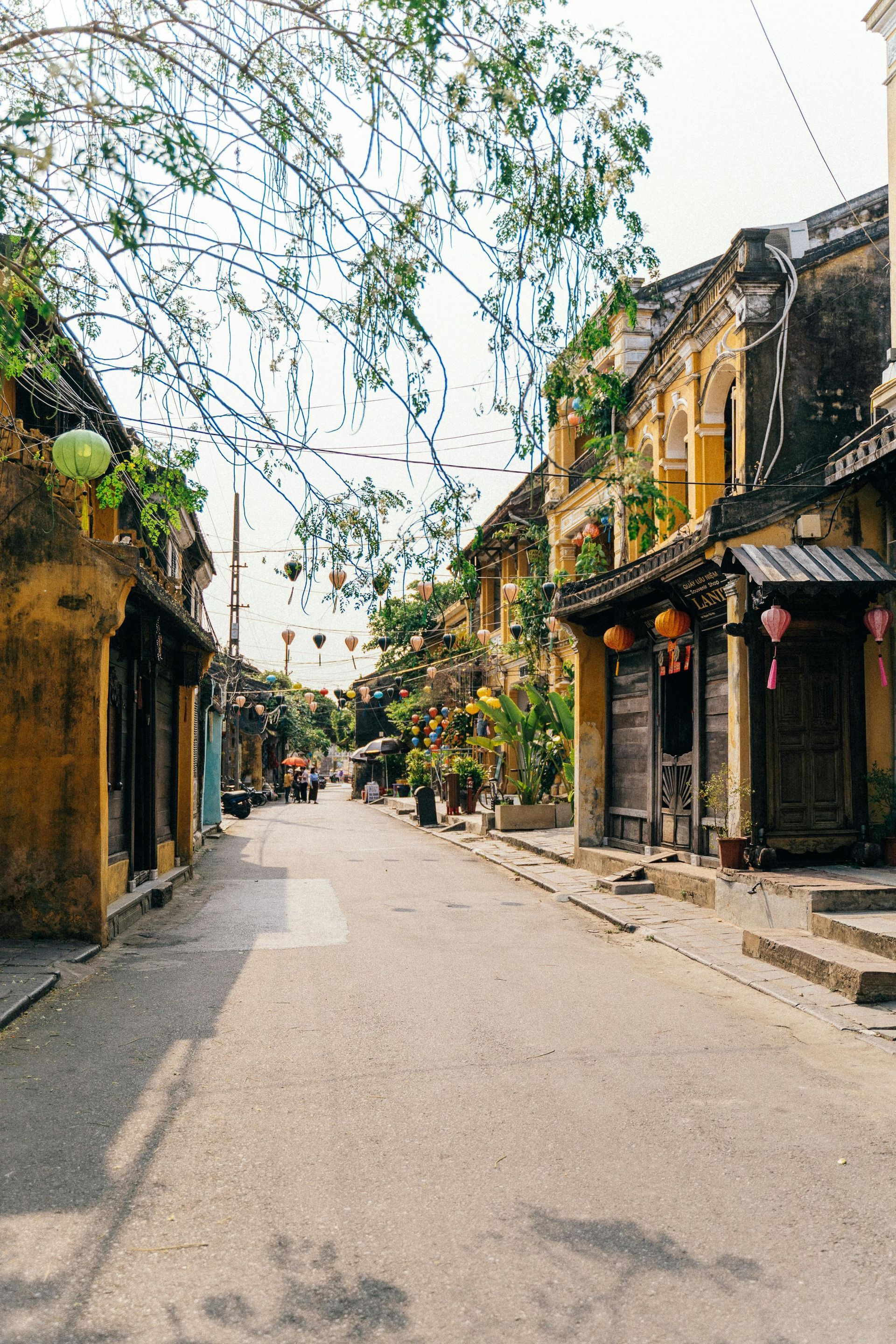 A narrow street lined with buildings and lanterns on a sunny day in Hoi An, Vietnam.