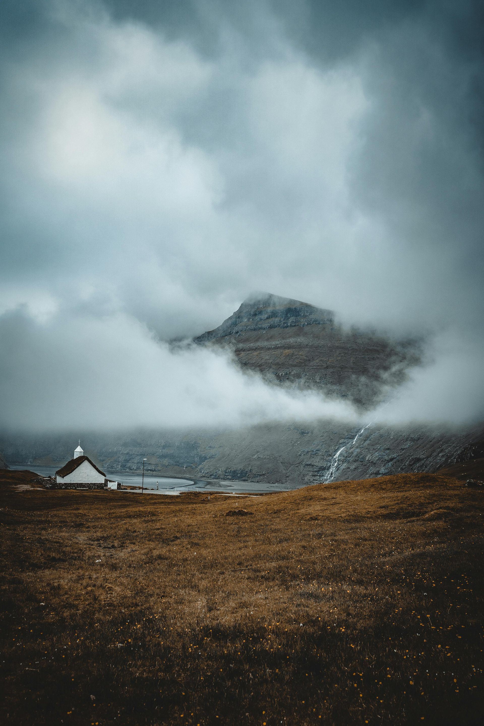 A small house in the middle of a field with a mountain in the background covered in clouds on the Faroe Islands.