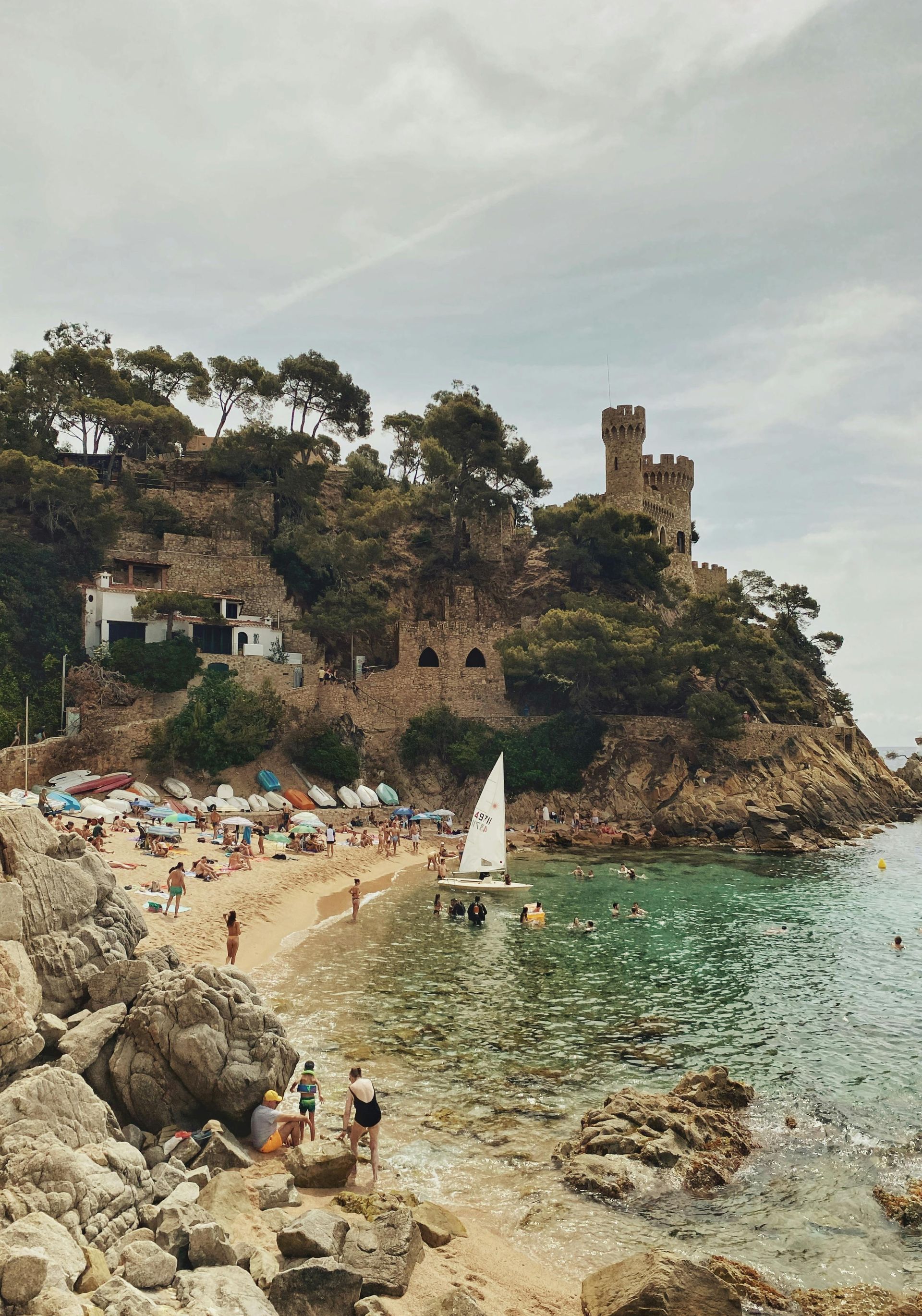 A sailboat is docked at a beach with a castle in the background on Costa Brava, Spain.