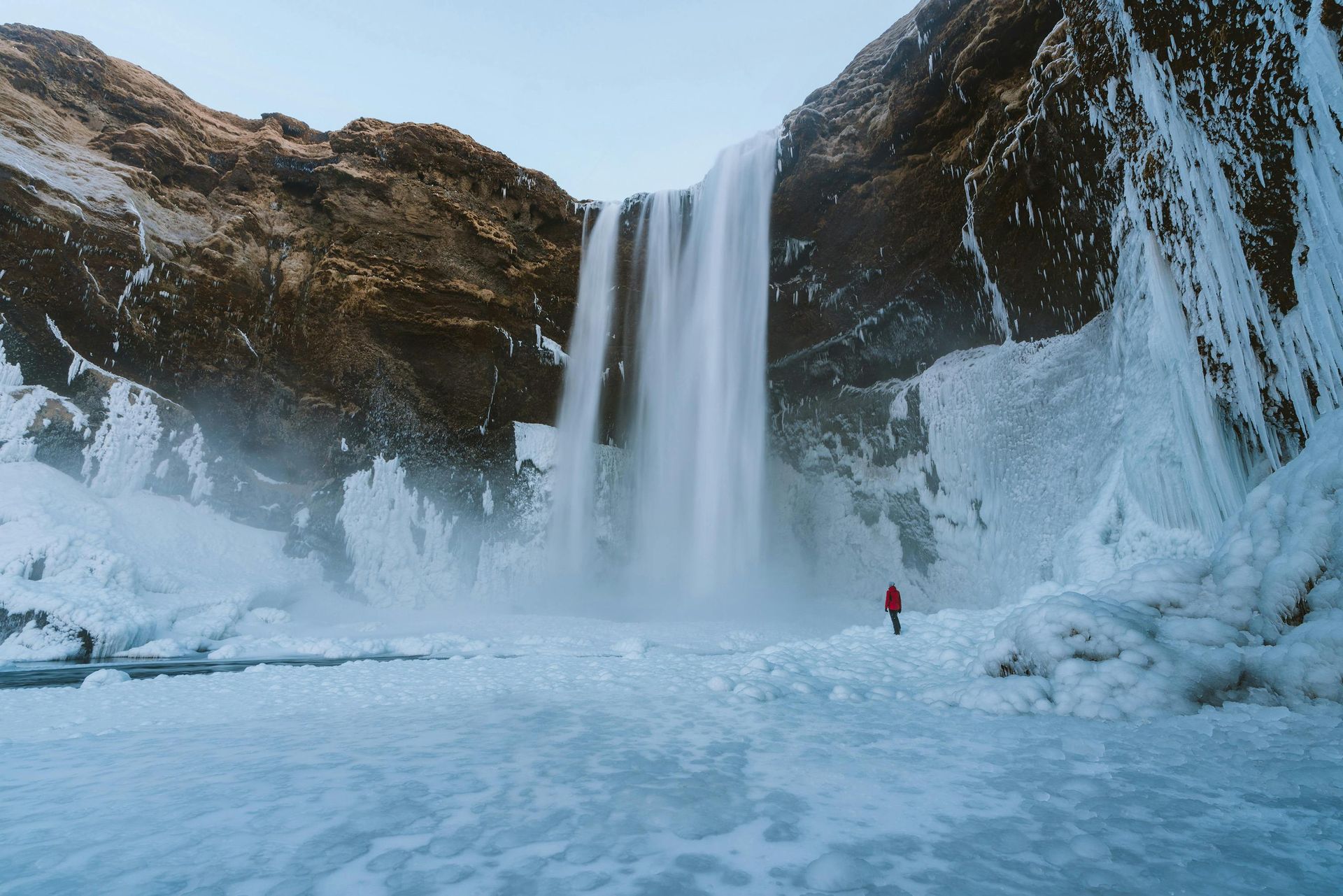 A person in a red jacket is standing in front of a waterfall in Iceland.