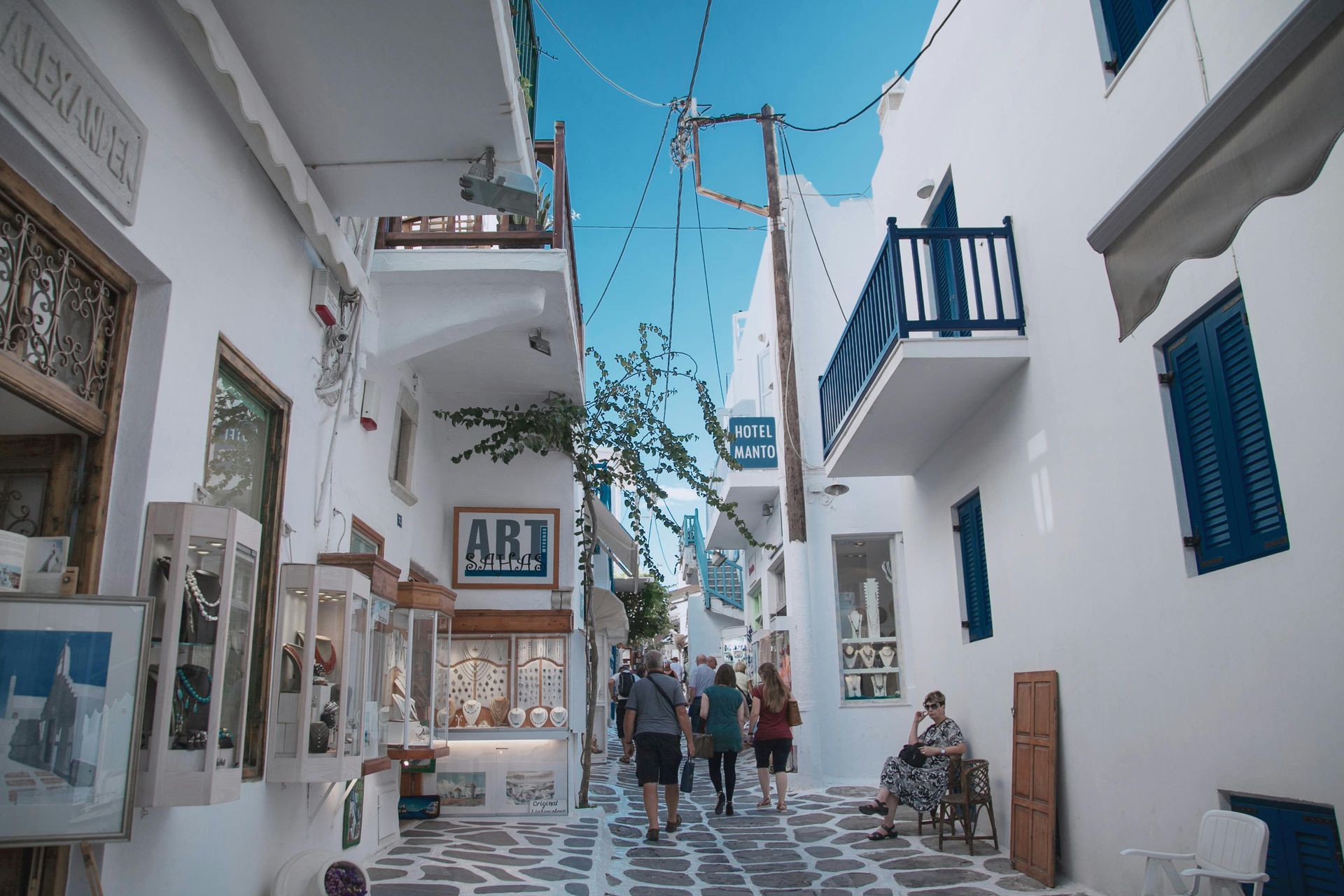 A group of people are walking down a narrow street between two buildings in Mykonos, Greece.