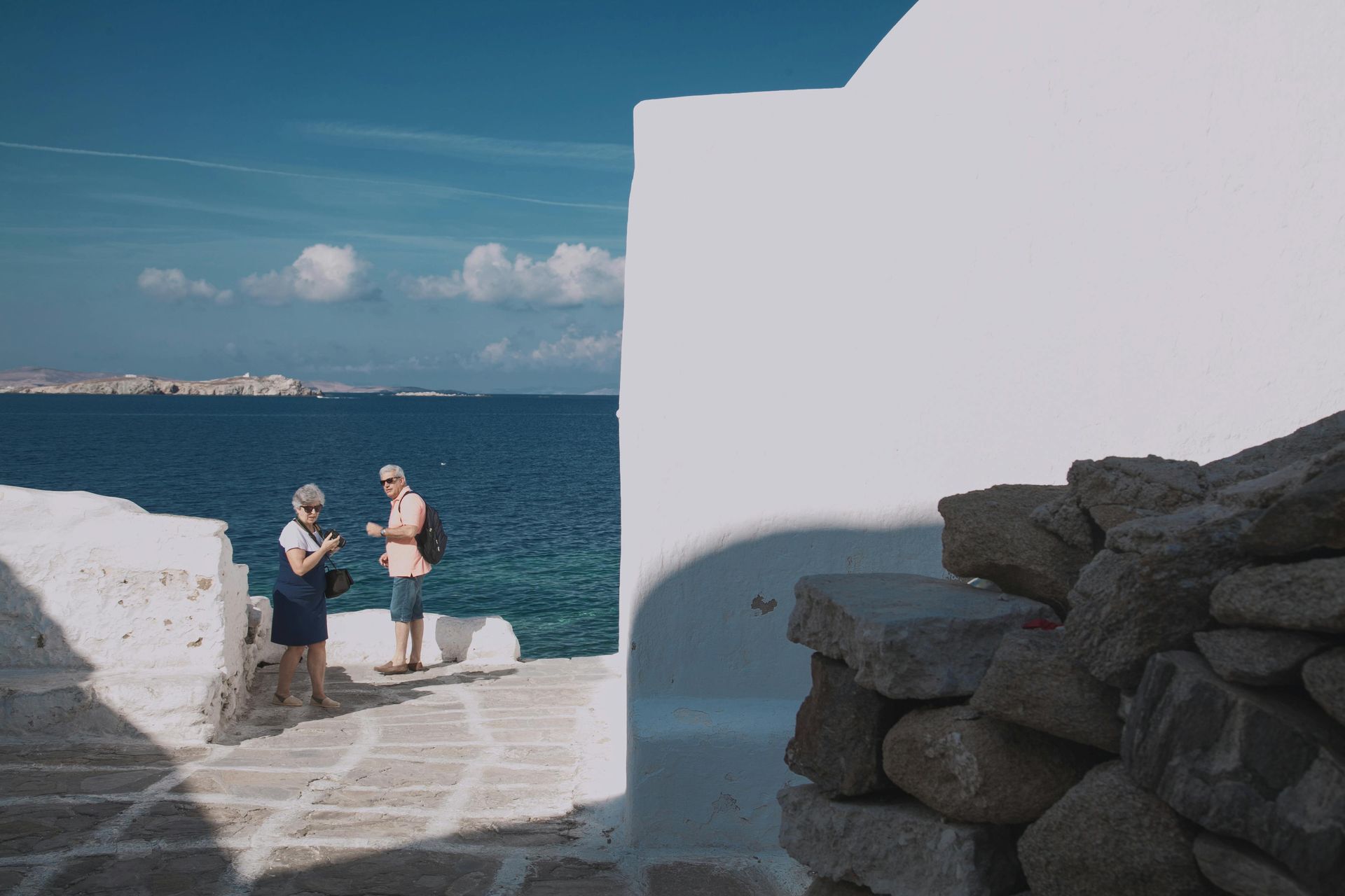 Two people are standing next to each other in front of a white building overlooking the ocean in Mykonos, Greece.