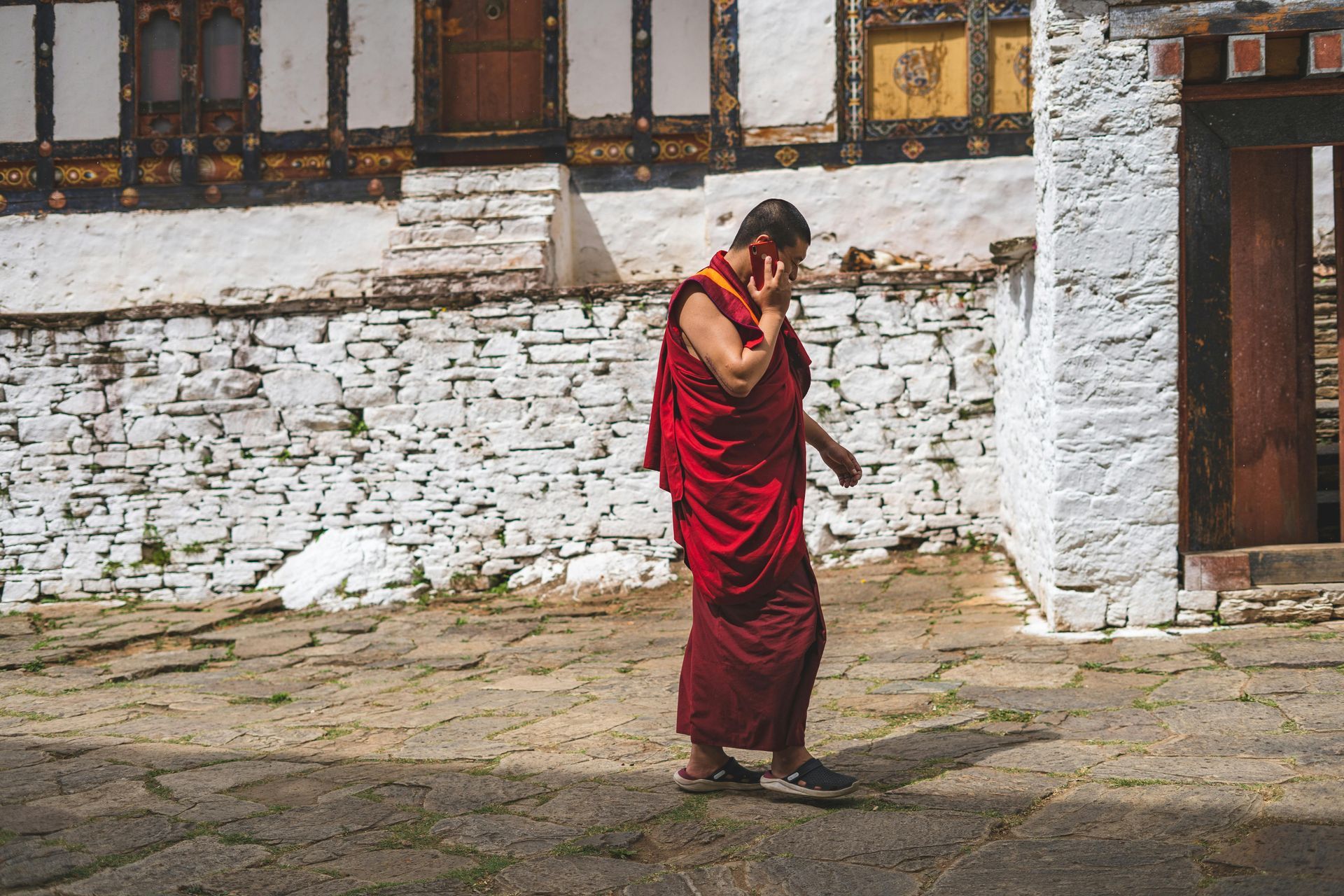 Monk in red robes talking on a cell phone near a white stone building in Paro Taktsang, Bhutan.