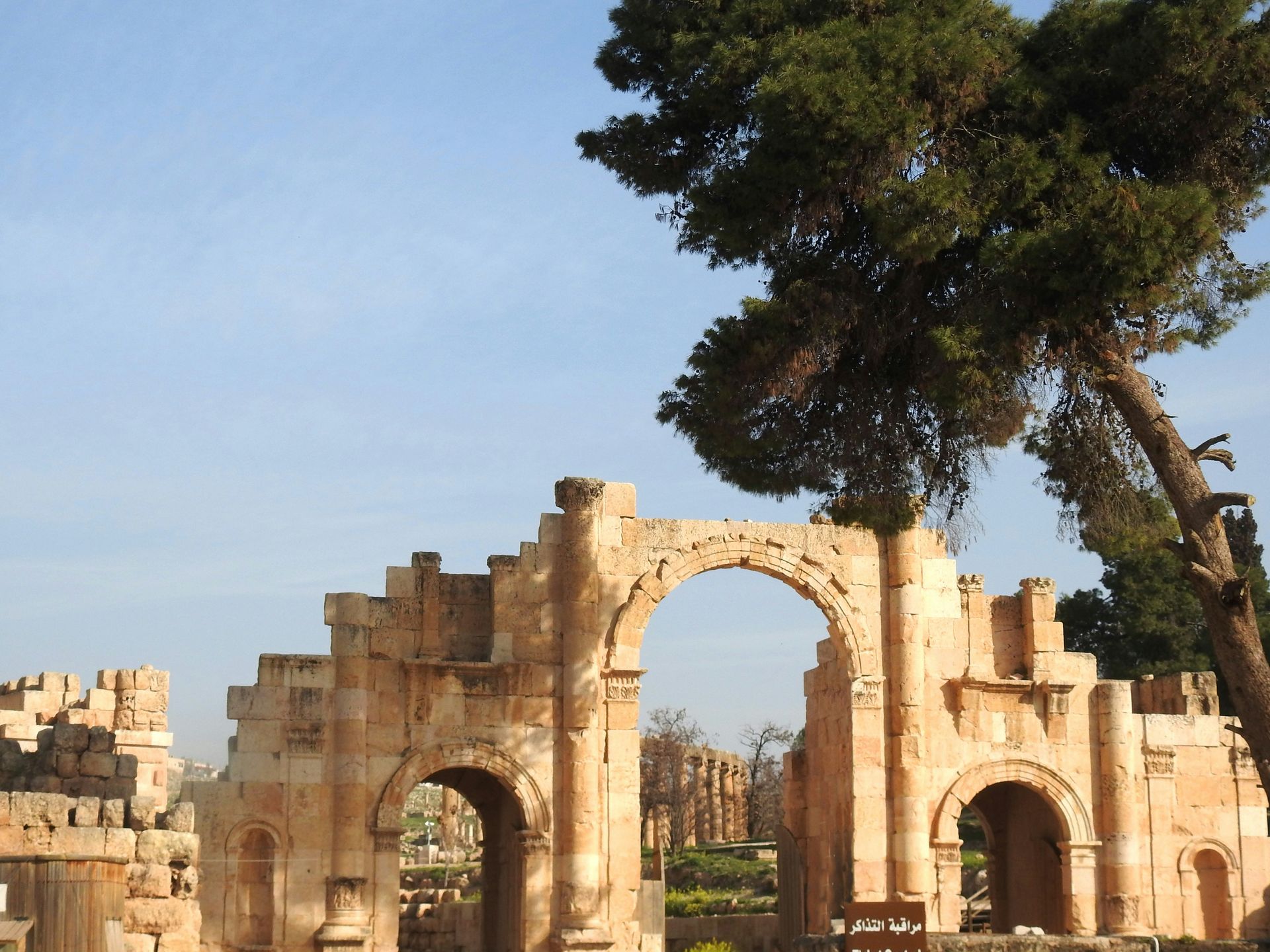 Ruins of an ancient Roman archway in Jerash, Jordan, with a tree in the background under a blue sky.