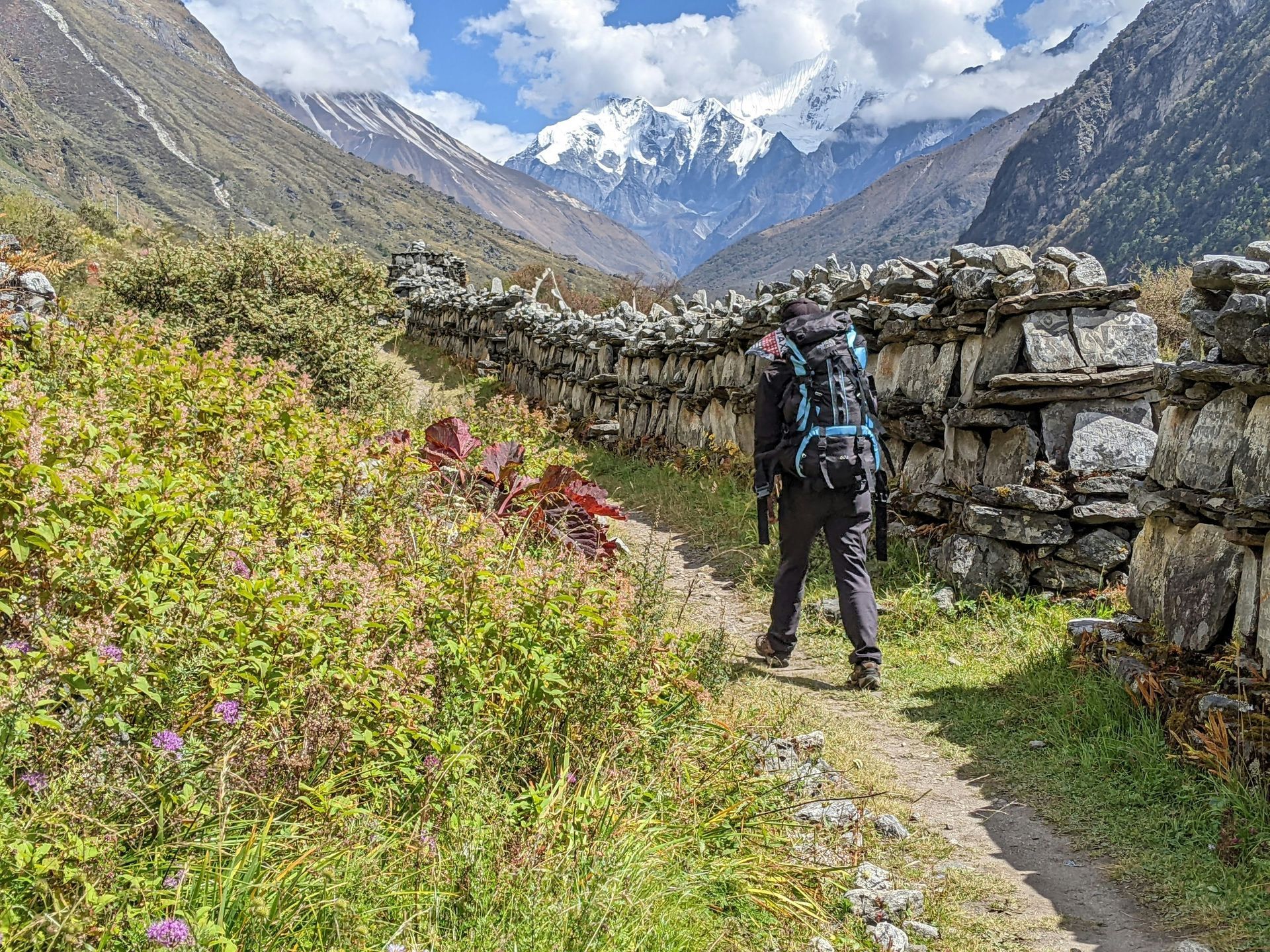 Person hiking on a trail next to a stone wall, mountains in Langtang, Nepal.