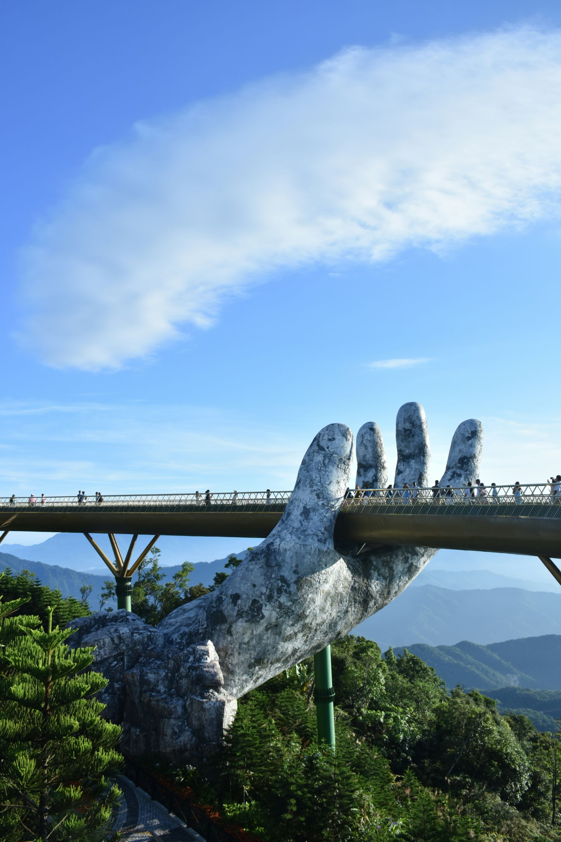 Golden Bridge supported by giant stone hands in Bà Nà Hills, Vietnam, with blue sky and tourists walking.