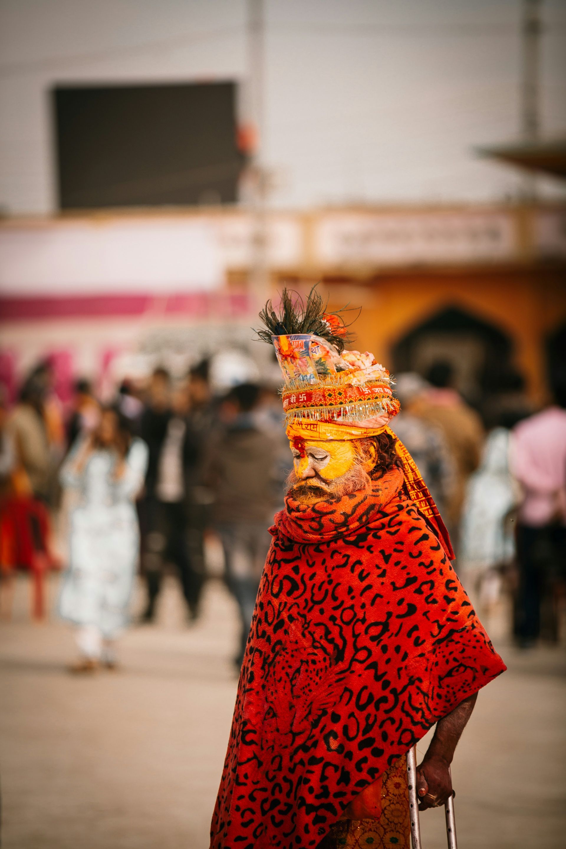 Man in orange and red robes with ornate headgear and face paint, holding a cane in Janakpur, Nepal.