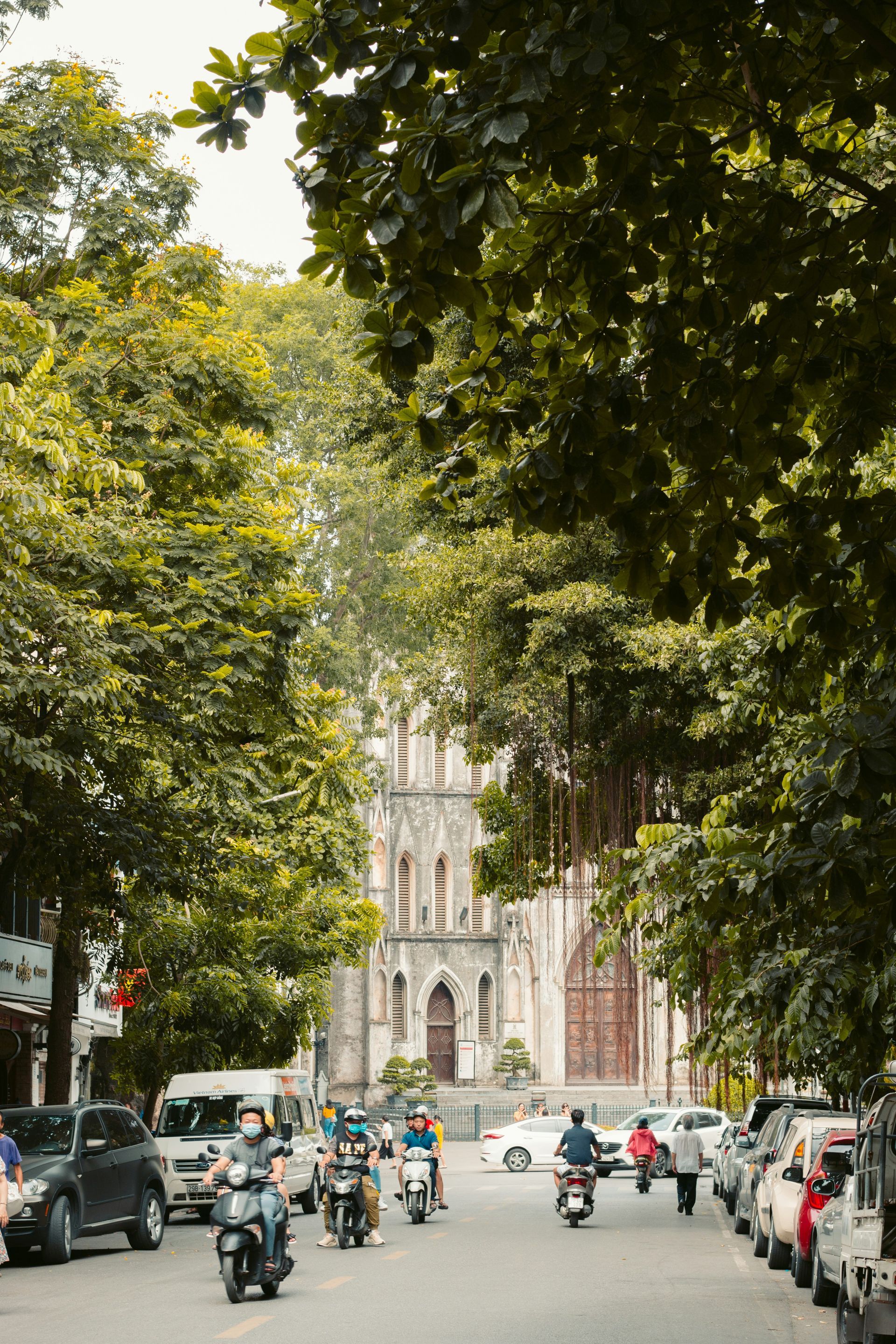 Street scene in Hanoi, Vietnam, with motorbikes, cars, lush trees, and a church in the distance.