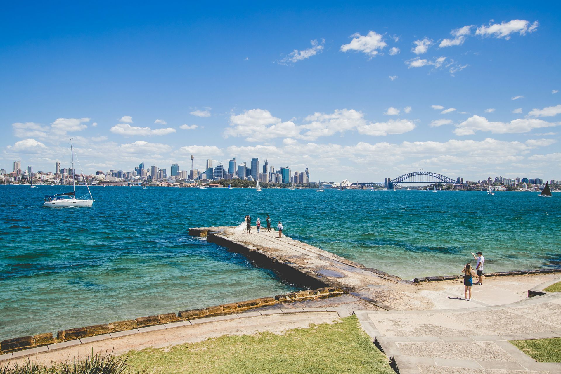 A group of people standing on a pier overlooking a body of water with the Sydney, Australia, skyline in the background.