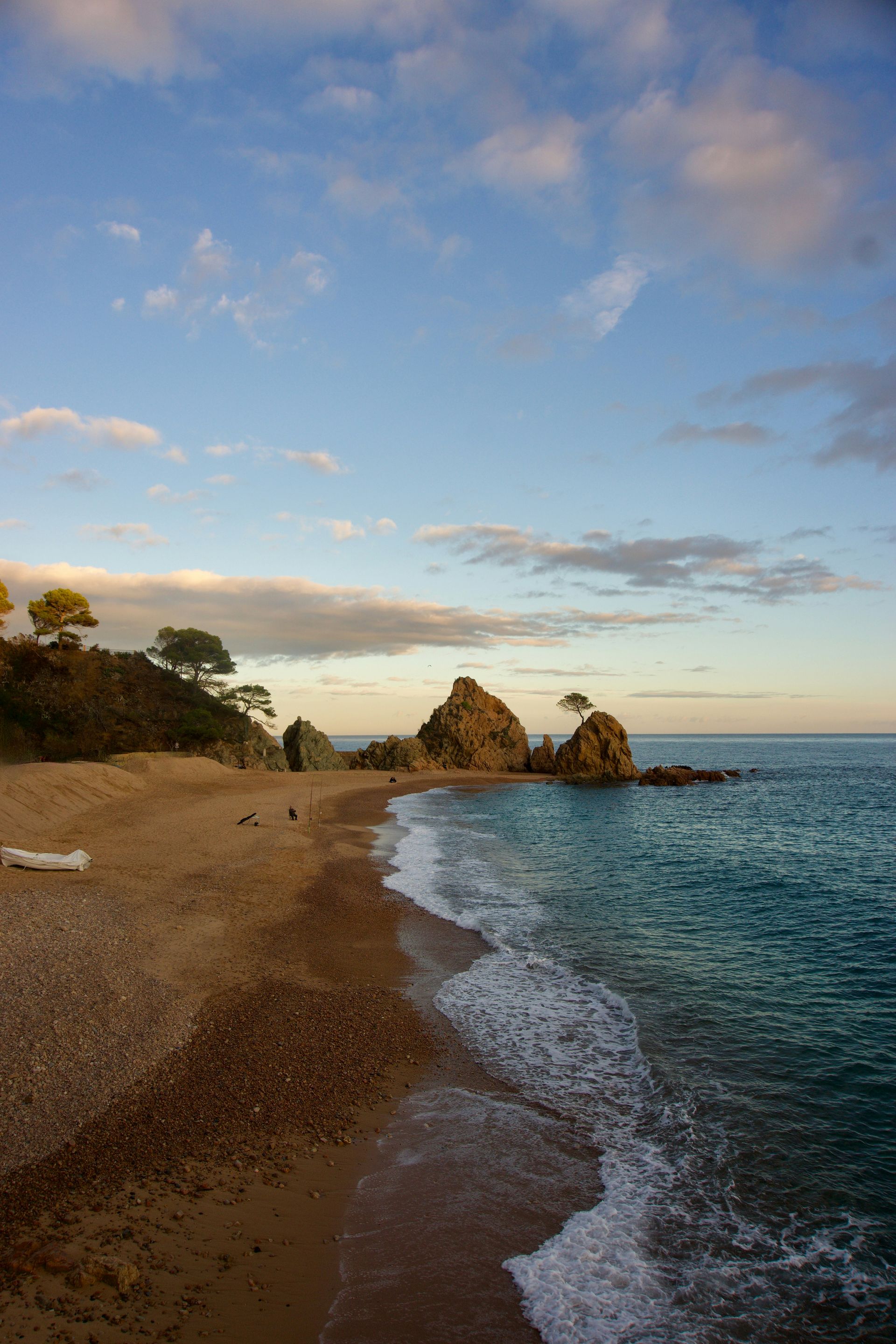 Beach with rocky shore and turquoise water under a partly cloudy blue sky on Costa Brava, Spain.
