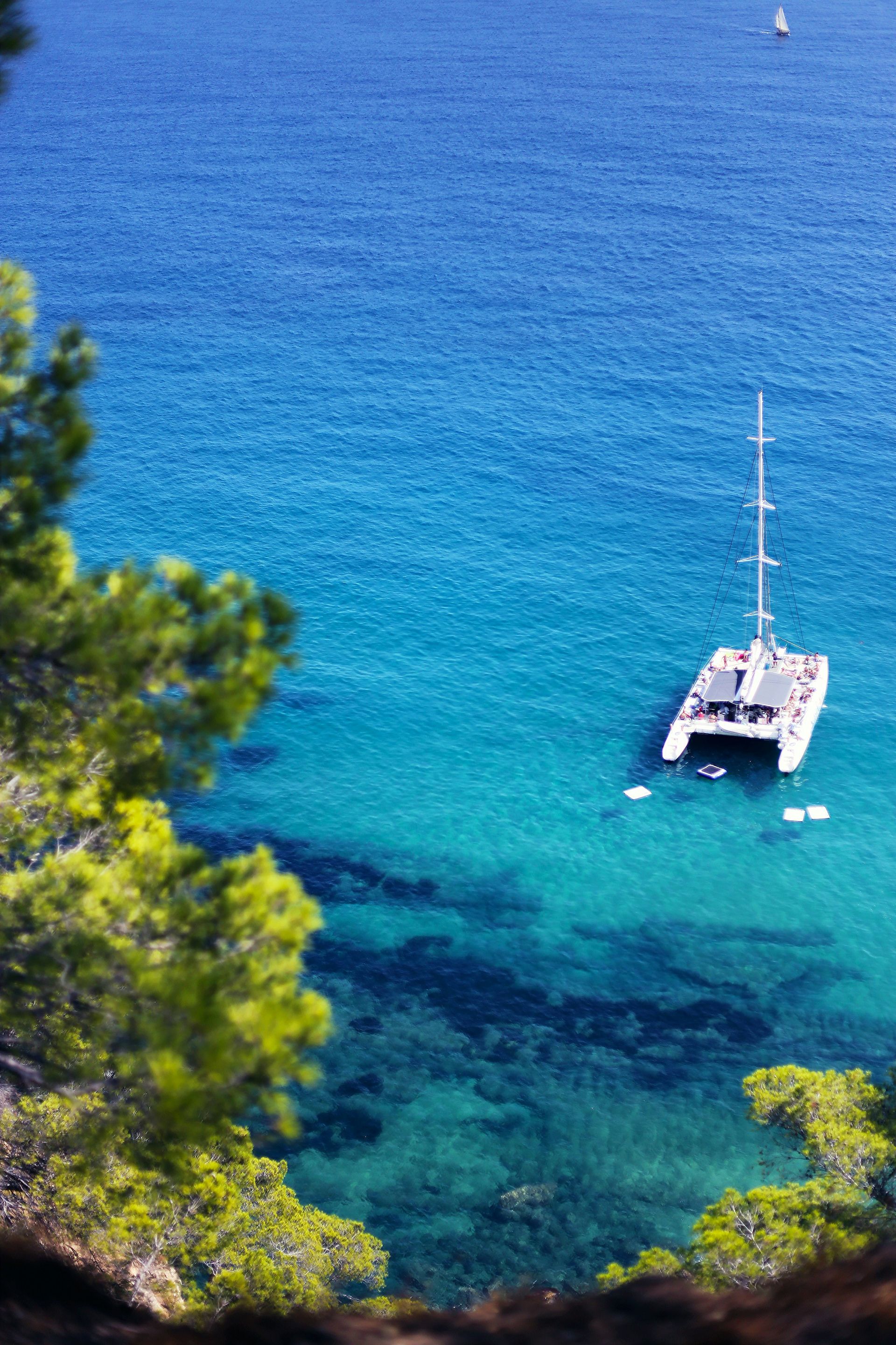 Catamaran anchored in turquoise water near a shoreline with pine trees on Costa Brava, Spain.