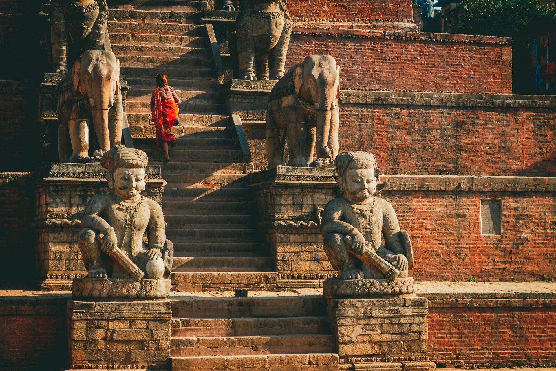 Stone statues flank a staircase with a person in red climbing it at a temple in Bhaktapur, Nepal.