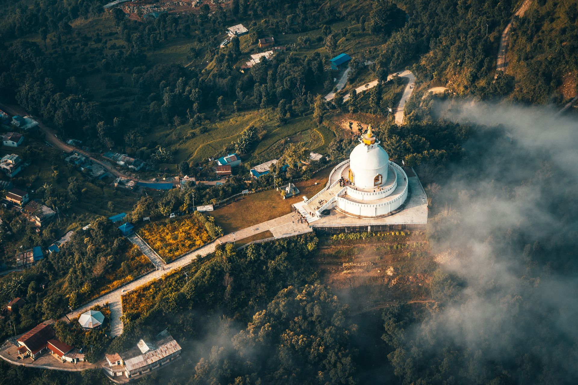 White Buddhist stupa atop a hill surrounded by lush green trees, and a winding road in Pokhara, Nepal.