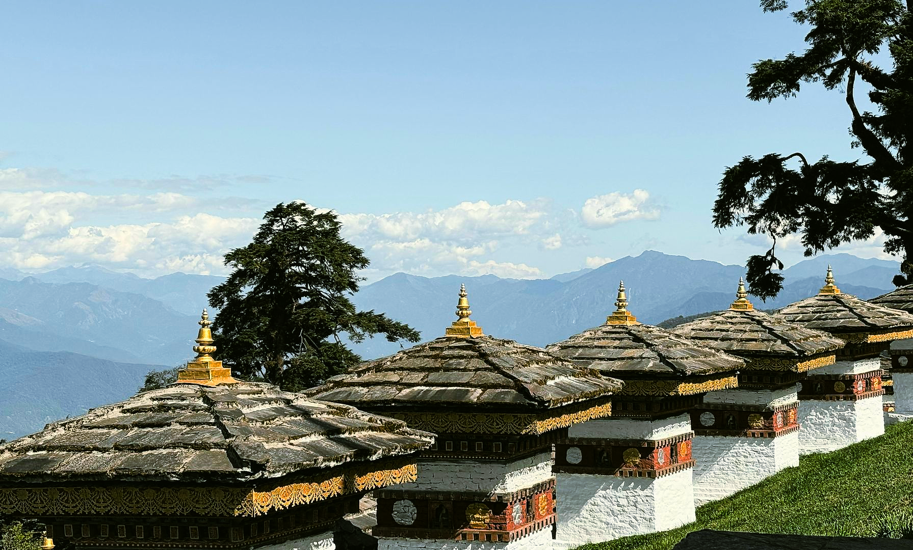 Row of white and gold Buddhist stupas on a grassy hill overlooking mountains at Dochula Pass, Bhutan.