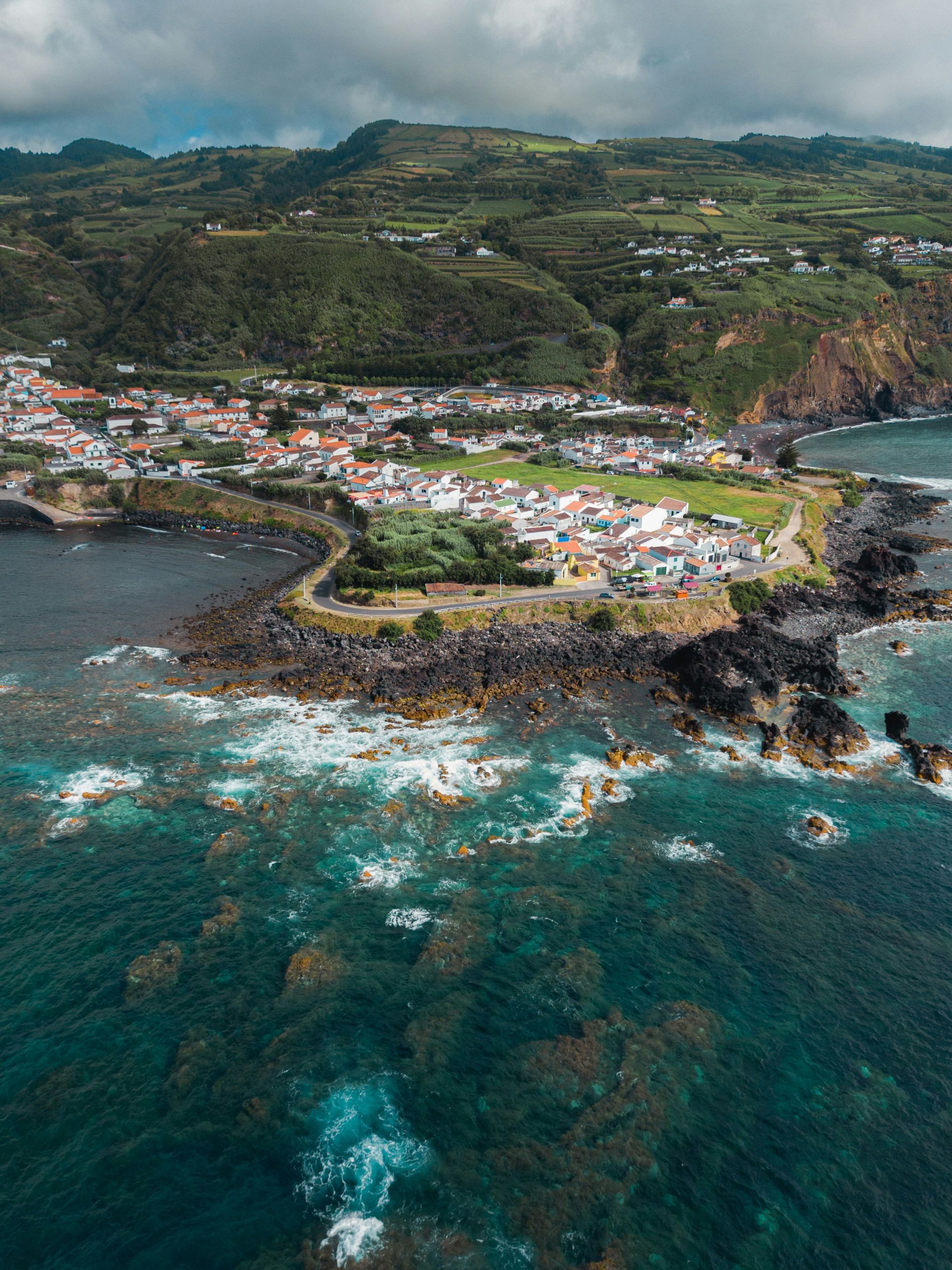 Coastal town nestled between rocky shore and green hills, with turquoise ocean on the island of Azores, Portugal.