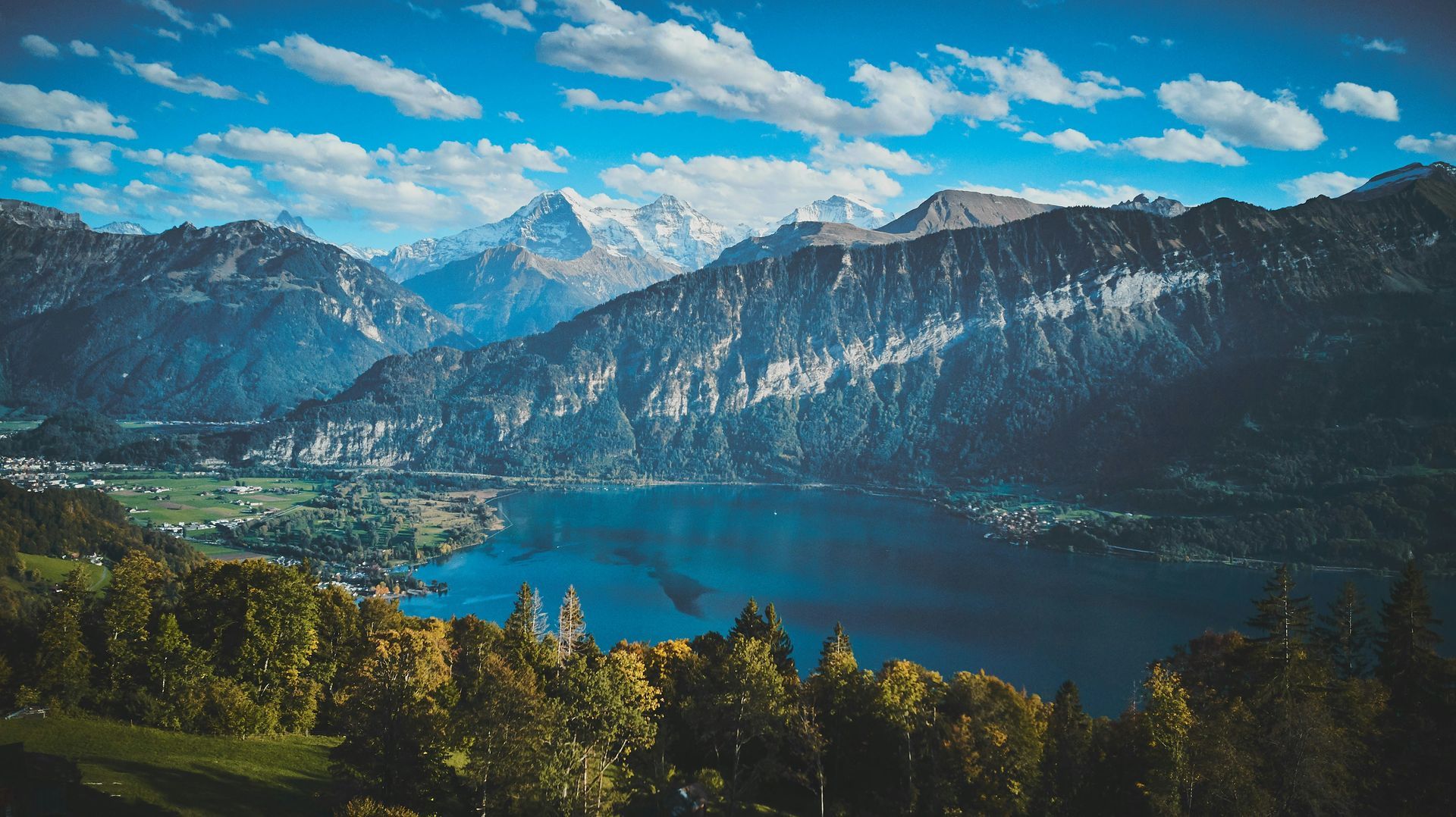 Mountains frame a blue lake with towns and trees. Snow-capped peaks under a cloudy sky in Interlaken, Switzerland.