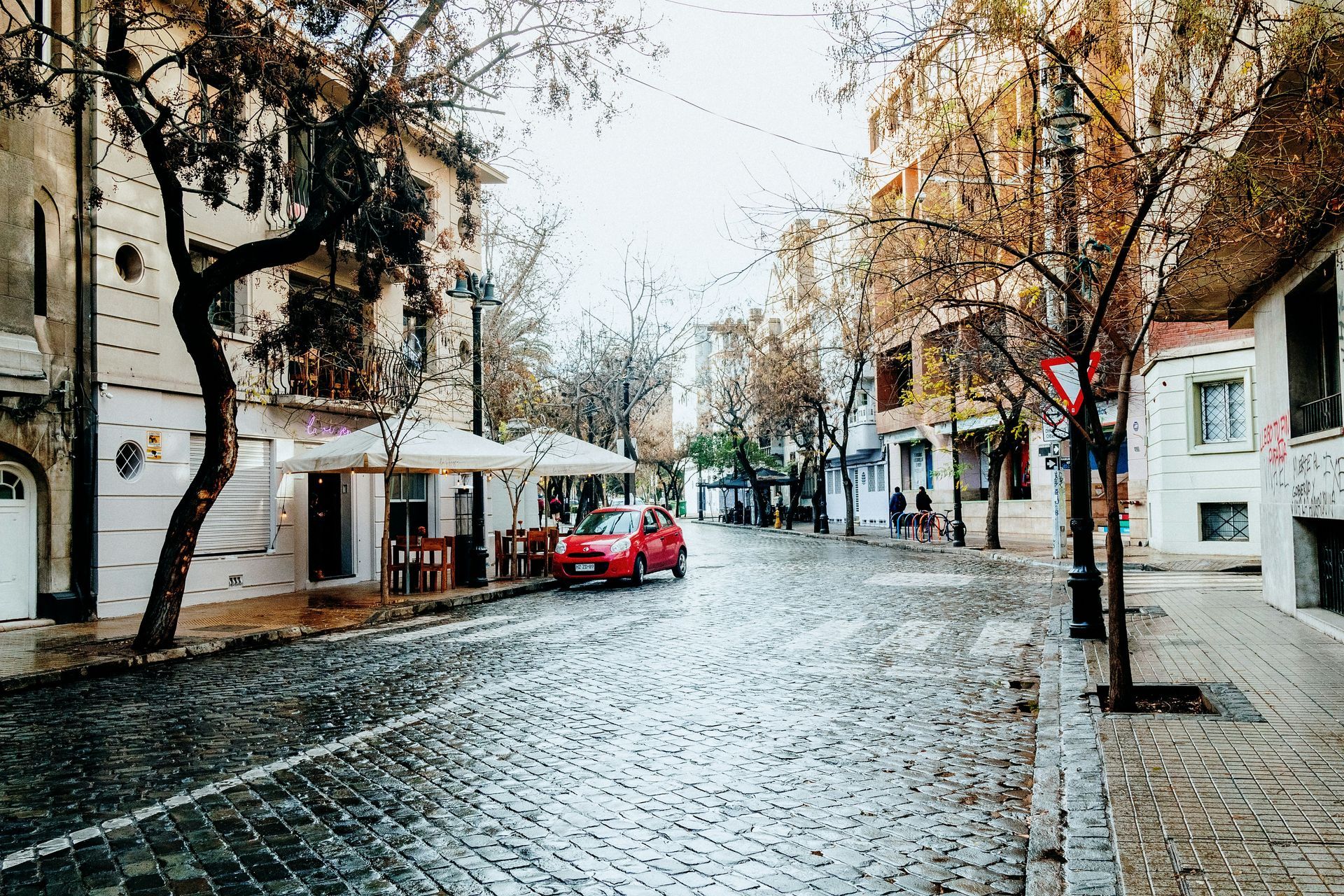 Cobblestone street with buildings, bare trees, and a red car in Santiago, Chile.