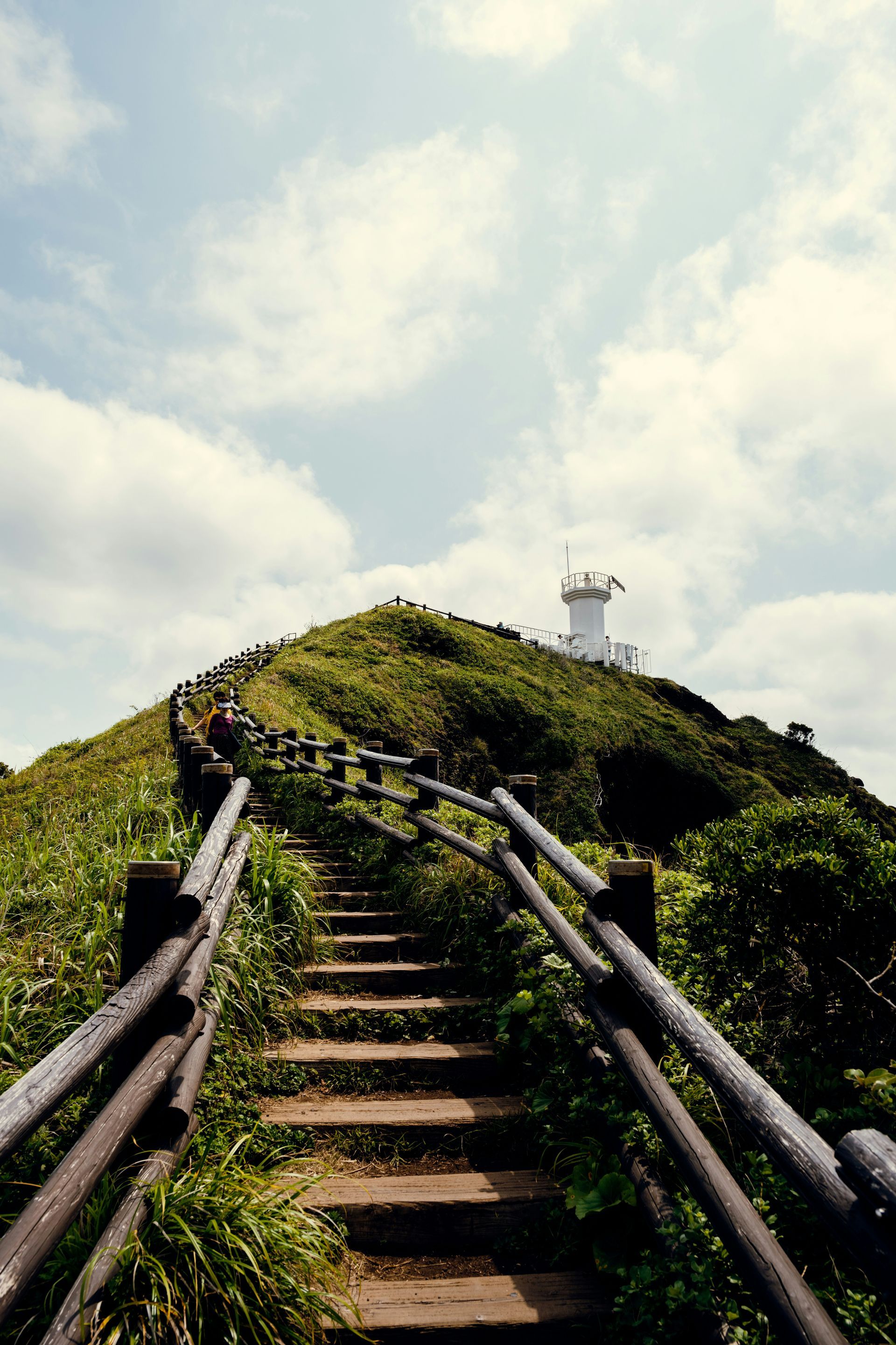 Stone stairs ascend a grassy hill to a white tower, wooden railings on either side on Jeju Island, South Korea.