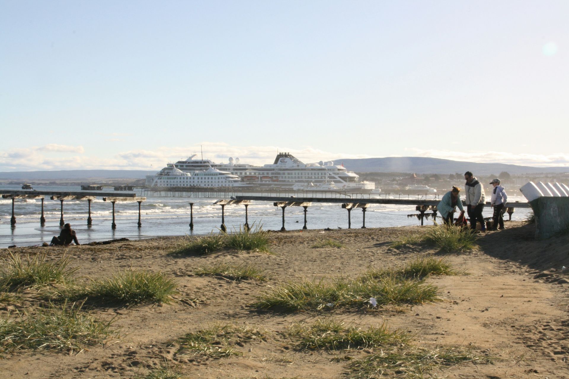 Beach scene: Pier with people, a large cruise ship in the distance, clear sky in Punta Arenas, Chile.