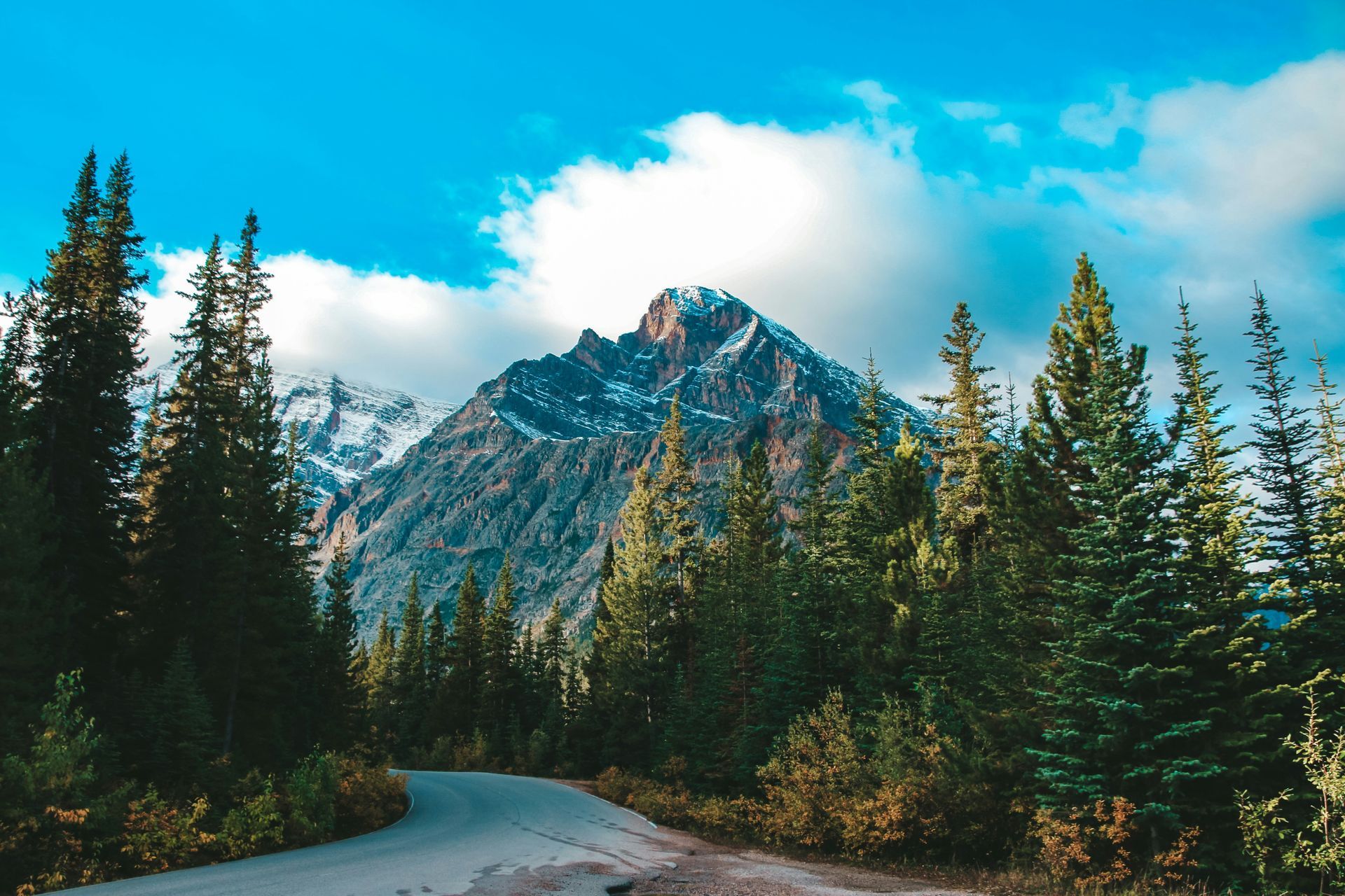 Road leading into a mountain range with snow-capped peak, surrounded by evergreen trees, in Banff, Canada.