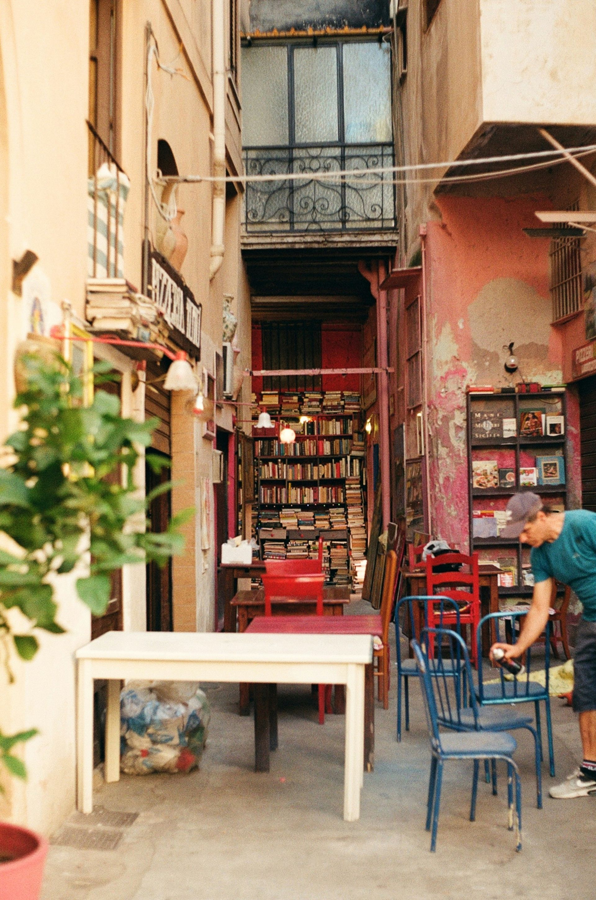 A man is standing in front of a bookstore with tables and chairs in Sicily.