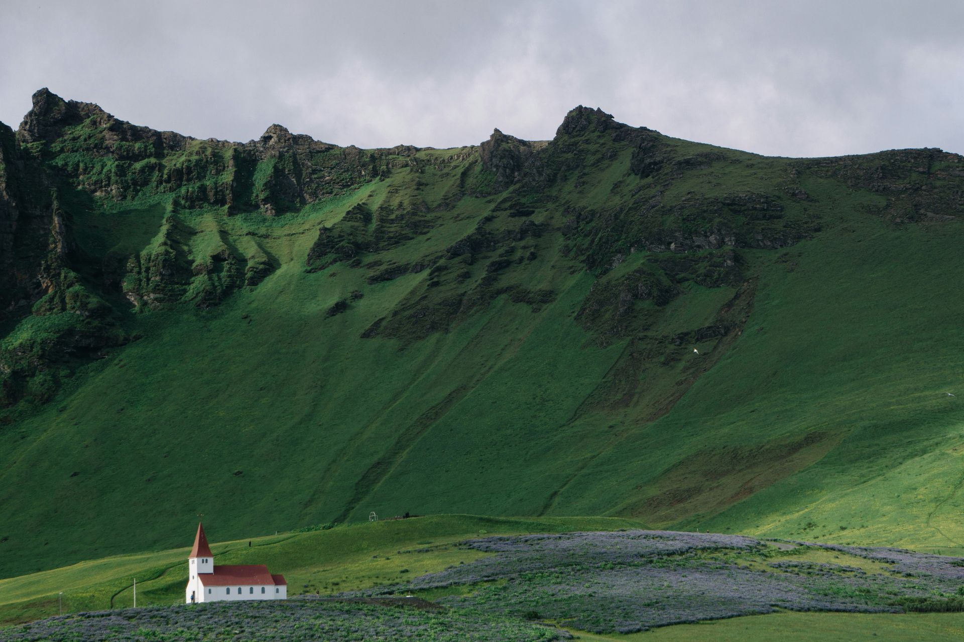 Church with red roof nestled in a green field against a rugged, mossy mountain in Vis, Iceland.