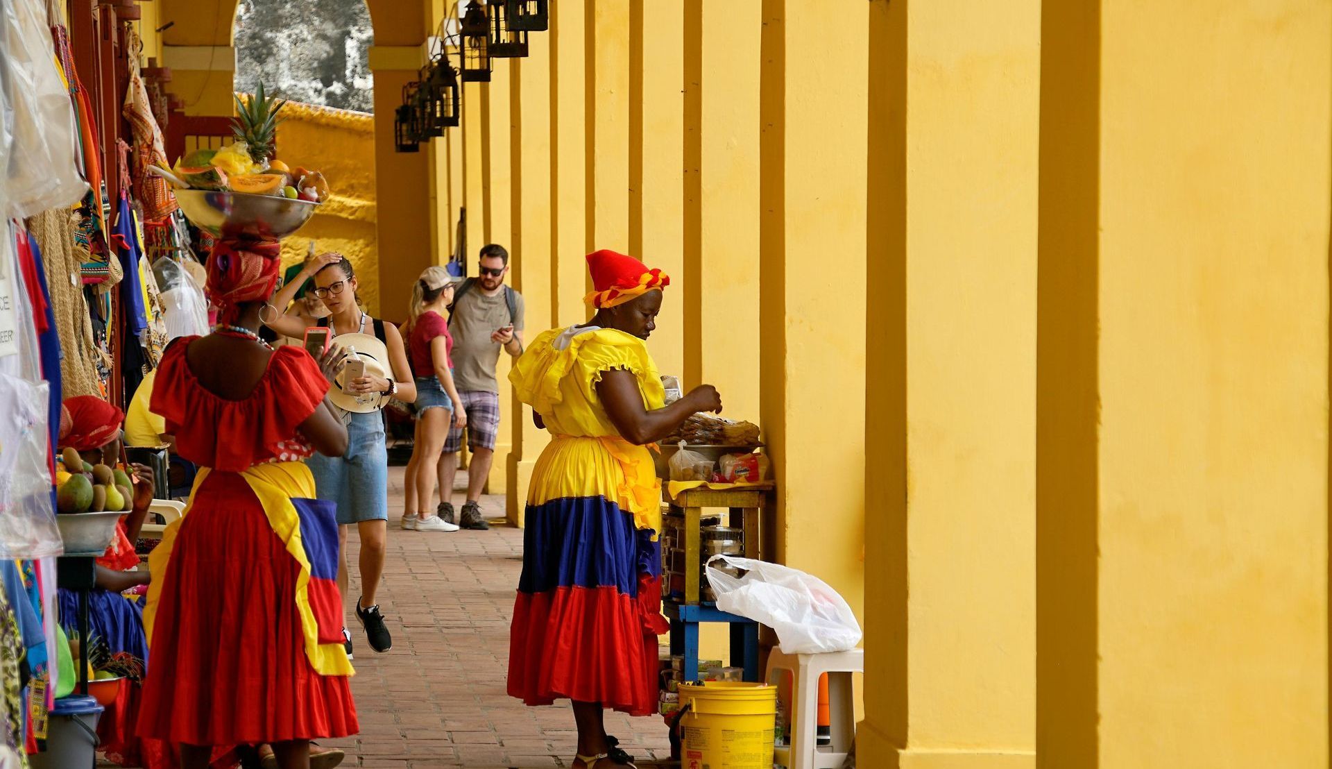 A woman in a red , yellow and blue dress is standing in front of a yellow building in Cartagena, Colombia.