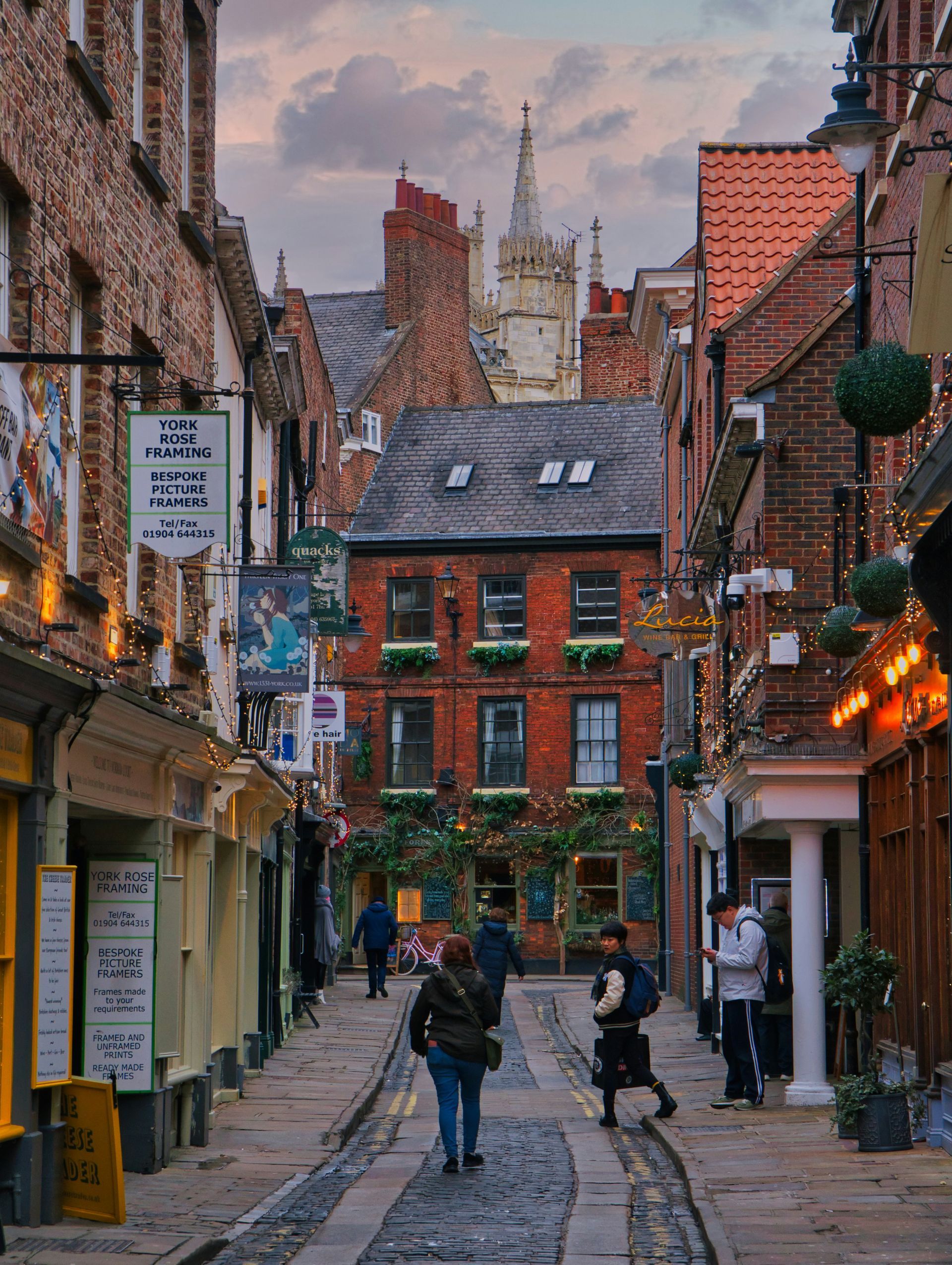 Narrow cobblestone street lined with brick buildings. A church spire rises in the distance in York, England.