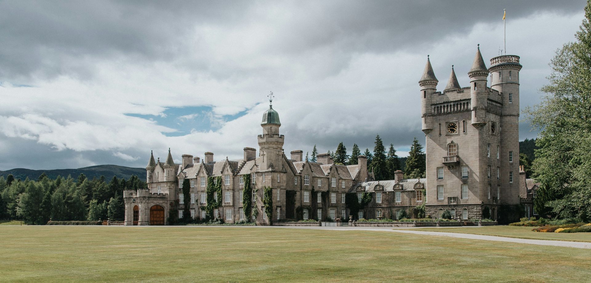 The Balmoral Castle, with multiple turrets and towers, is set in a green field under a cloudy sky in Scotland.