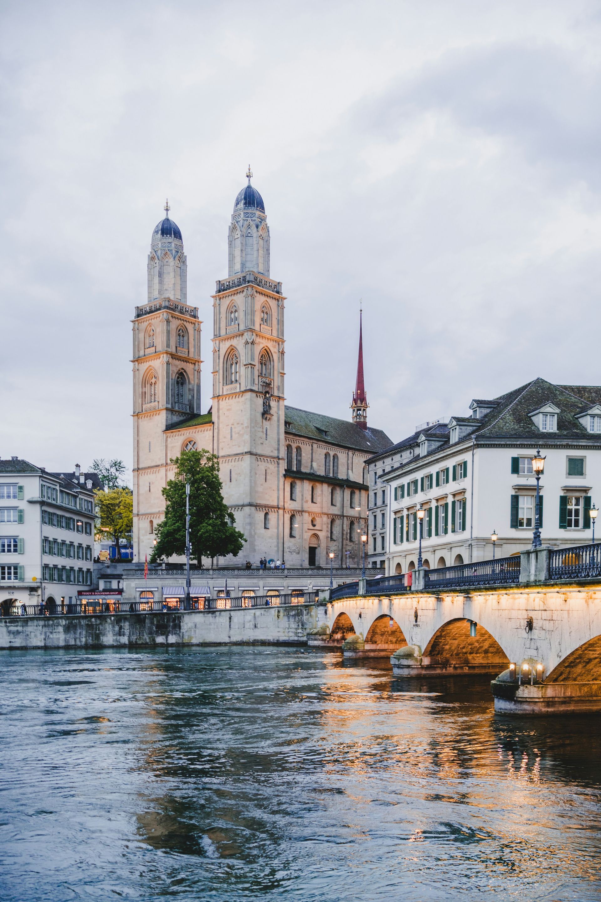 Twin-towered Grossmünster church in Zurich overlooking the Limmat river, with a bridge and buildings in the evening light.