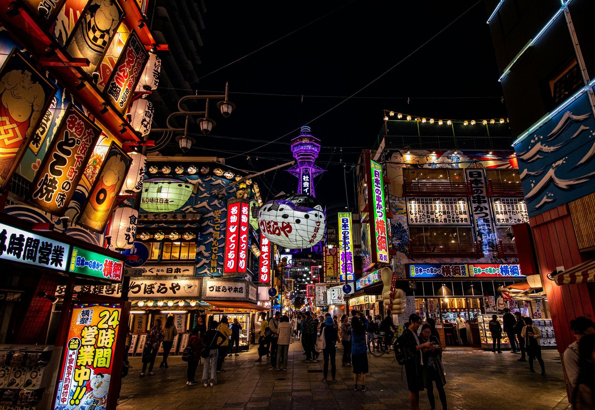 Nighttime view of a crowded street in Osaka, Japan, with bright neon signs and the Tsutenkaku Tower.