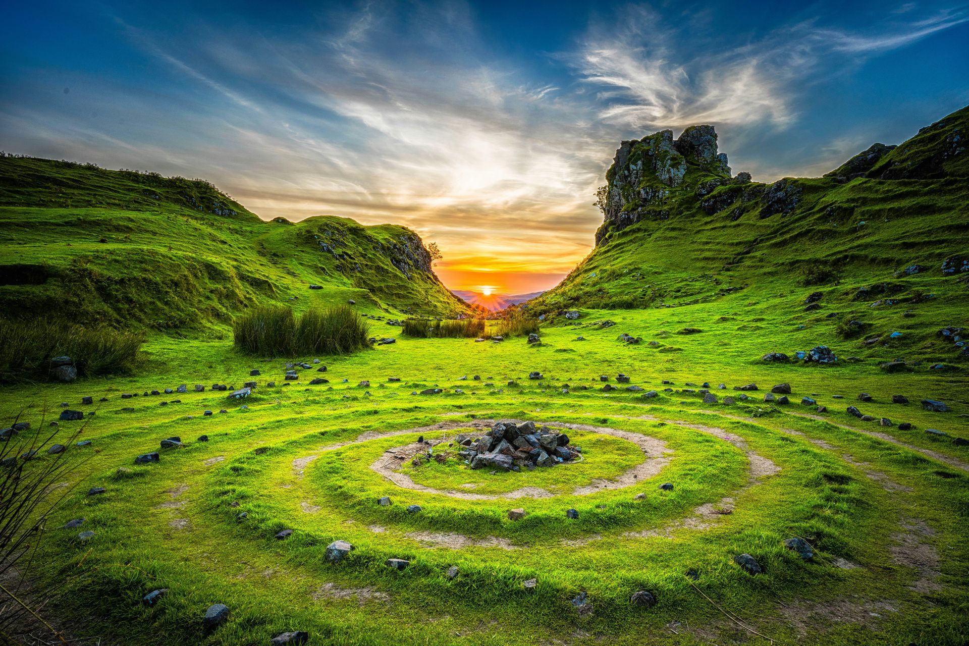 Rolling green hills with a stone spiral, bright sunset in the distance at the Fairy Glen in Scotland.